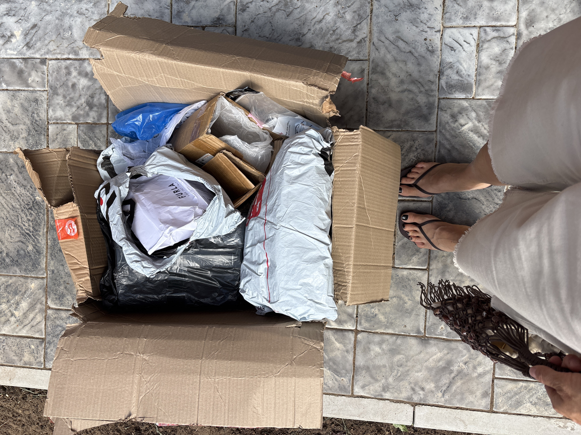 An eye-level shot depicts an open, partially torn cardboard box overflowing with packaging materials and objects, set on a gray, tiled surface. The packaging includes items wrapped in white and black plastic bags, smaller cardboard boxes, and blue plastic. A white piece of paper with the word "FURLA" is visible among the contents.
To the right, a person's lower legs and feet are seen. The person is wearing white clothing and black flip-flops with dark nail polish. They are holding a dark brown, macrame-style bag.
The scene appears to be outdoors on a patio or similar paved area. The location is the city of Chaikovsky, Russia.