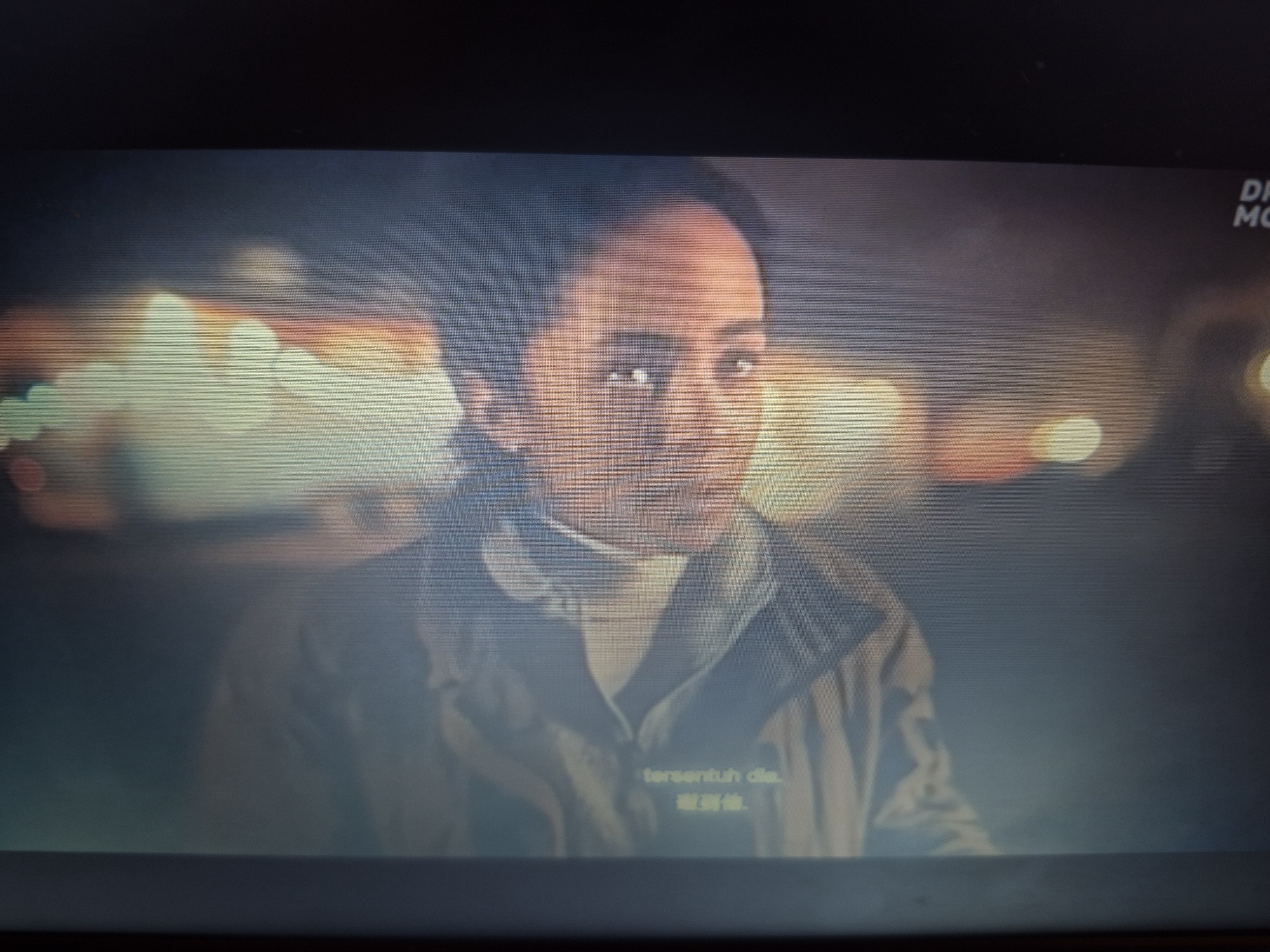 The image is a still from a video, likely a movie or television show, displaying a woman's face in close-up. She has dark hair and is looking directly at the camera with a serious expression. The lighting is dim and appears to be artificial, possibly from streetlights or vehicle headlights, creating a bokeh effect in the background with blurred lights. There is text overlaid on the lower portion of the image, in what appears to be a foreign language. The overall aesthetic suggests a night scene in an urban environment. There is no discernible information within the image to indicate a specific location within the city of Sandwell, United Kingdom.