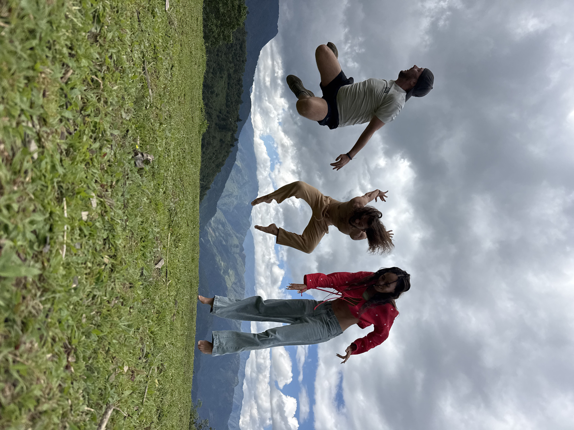 Three individuals are captured mid-air in a synchronized jump against a backdrop of mountains and a partly cloudy sky in Tú Lệ, Vietnam. The foreground consists of lush, short green grass.
From left to right:
A male individual with light skin, a short beard, and a dark baseball cap is seen jumping, his body angled right. He wears a light-colored short-sleeved shirt and dark shorts, with athletic shoes on his feet. His arms are bent, and his legs are tucked.
In the center, another individual, appearing male, with long, wavy brown hair, is shirtless and wearing light brown pants. This person is also mid-jump, with arms raised and hands open in a dynamic, dance-like pose. Their feet are bare.
On the right, a female individual with braided dark hair is jumping. She wears a bright red long-sleeved crop top adorned with small white heart patterns, paired with light blue wide-leg jeans. Her arms are extended forward, hands open, and her feet are bare.
The background features several layers of green mountains under a sky that is predominantly covered with dark grey clouds, though patches of blue sky are visible to the right. The overall scene conveys a sense of joy and freedom in a natural, open environment.