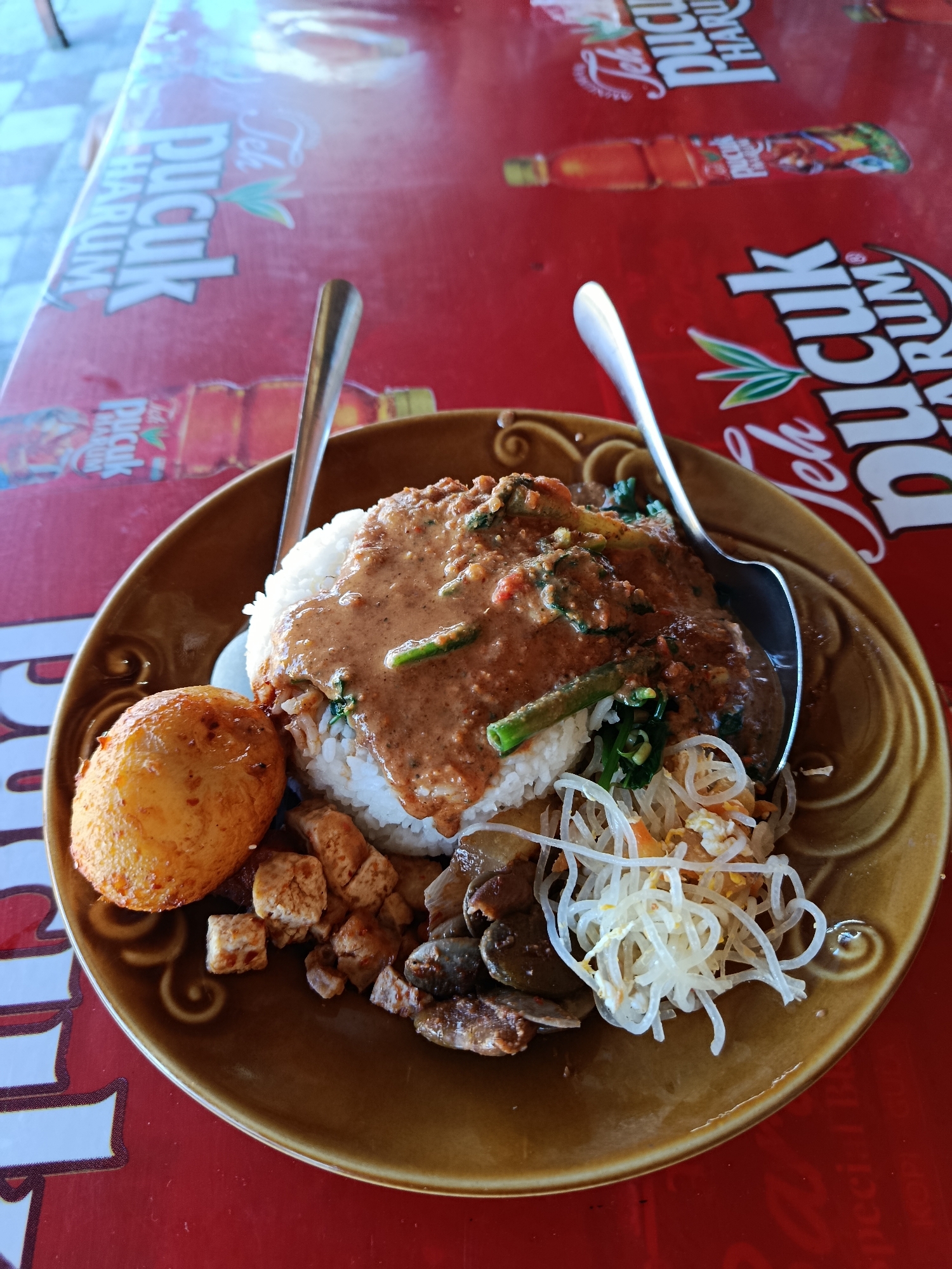 A close-up overhead shot captures a plate of Indonesian food, specifically Nasi Pecel, served on a round, brown ceramic plate with a swirled decorative edge. The dish consists of white rice topped with a generous portion of peanut sauce, mixed with vegetables such as long beans and leafy greens. To the left of the rice is a whole, fried egg with a golden-brown, textured coating. Below the rice and egg are several small cubes of fried tofu, some chopped liver or gizzards, and a pile of thin, white rice vermicelli noodles mixed with shredded carrots and scrambled egg. The plate is placed on a red table with a repeating graphic pattern of "Teh Pucuk Harum," a brand of Indonesian bottled tea, featuring stylized green leaves and red bottle silhouettes. Two spoons are visible, one larger spoon resting partially on the plate, and a smaller spoon to the left. The lighting suggests an outdoor or well-lit indoor setting, casting soft shadows. The background is slightly blurred, but appears to be an outdoor environment with hints of a light-colored floor. The presence of the "Teh Pucuk Harum" branding strongly suggests the location is likely a restaurant or food stall in Indonesia, possibly Cemorolawang, given its common association with tourist eateries.