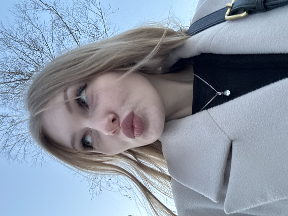 An adult female is depicted outdoors under a clear light blue sky in the city of Perm, Russia. Her light-colored hair, appearing blonde or light brown, is visible around her face, which is turned slightly upwards and to the left with her eyes looking in the same direction. Her lips are pursed in a "kissing" expression. She wears a light-colored, possibly beige or off-white, collared coat over a dark, likely black, garment. A silver-colored chain with a small, clear, teardrop-shaped pendant rests on her dark top. A dark strap with a gold-colored buckle is visible on her right shoulder (viewer's left). Bare, leafless tree branches are silhouetted against the sky in the upper background, indicative of a colder season.