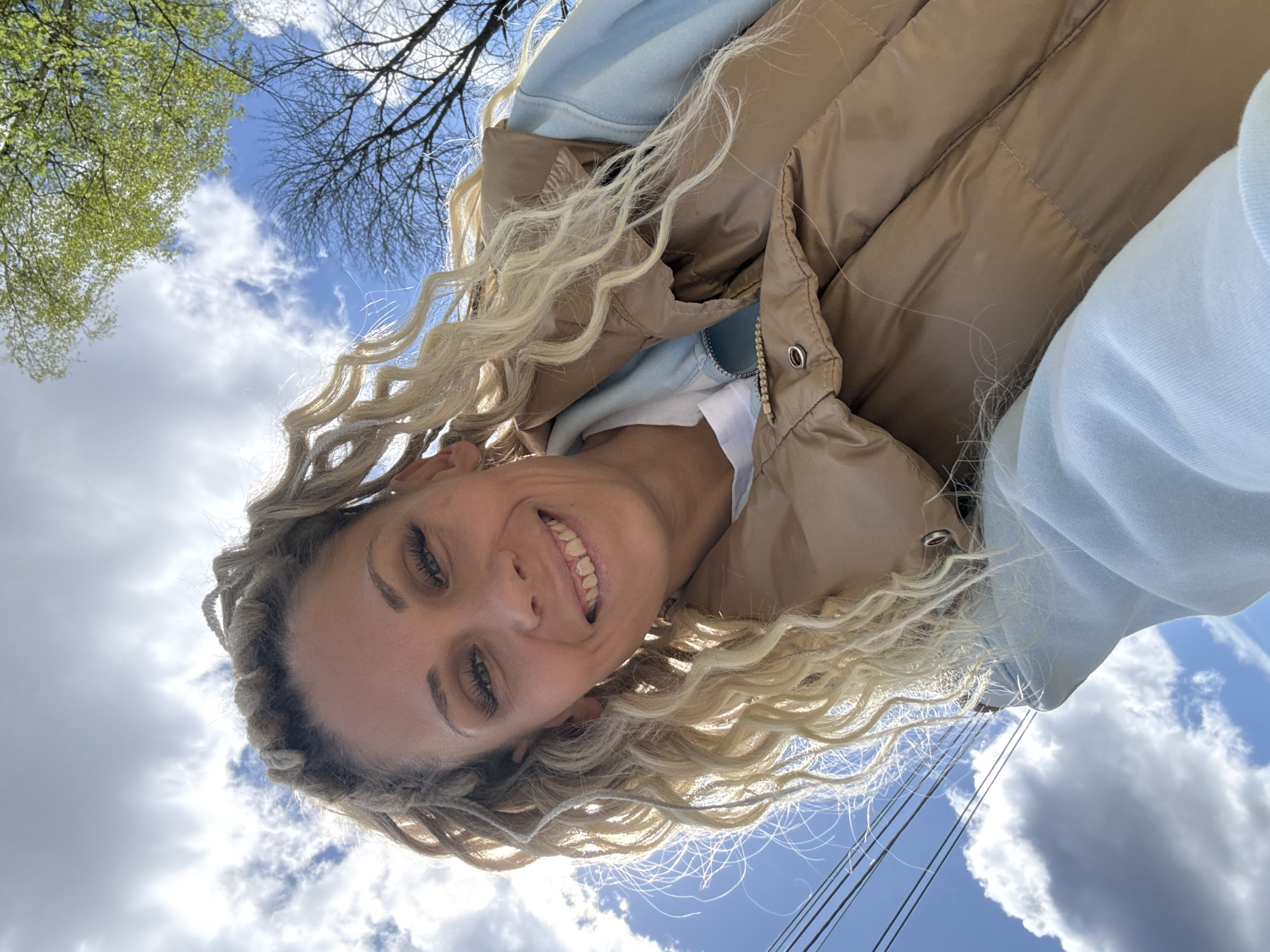 A woman with blonde, curly hair is seen from a low angle, likely a selfie, against a backdrop of a partly cloudy blue sky. She is wearing a light blue hoodie and a brown puffer vest. Power lines are visible in the lower right corner of the frame. Green leaves on tree branches can be seen at the top left of the frame. It is impossible to confirm that the picture was taken in the city of Rusanovo, Russia.
