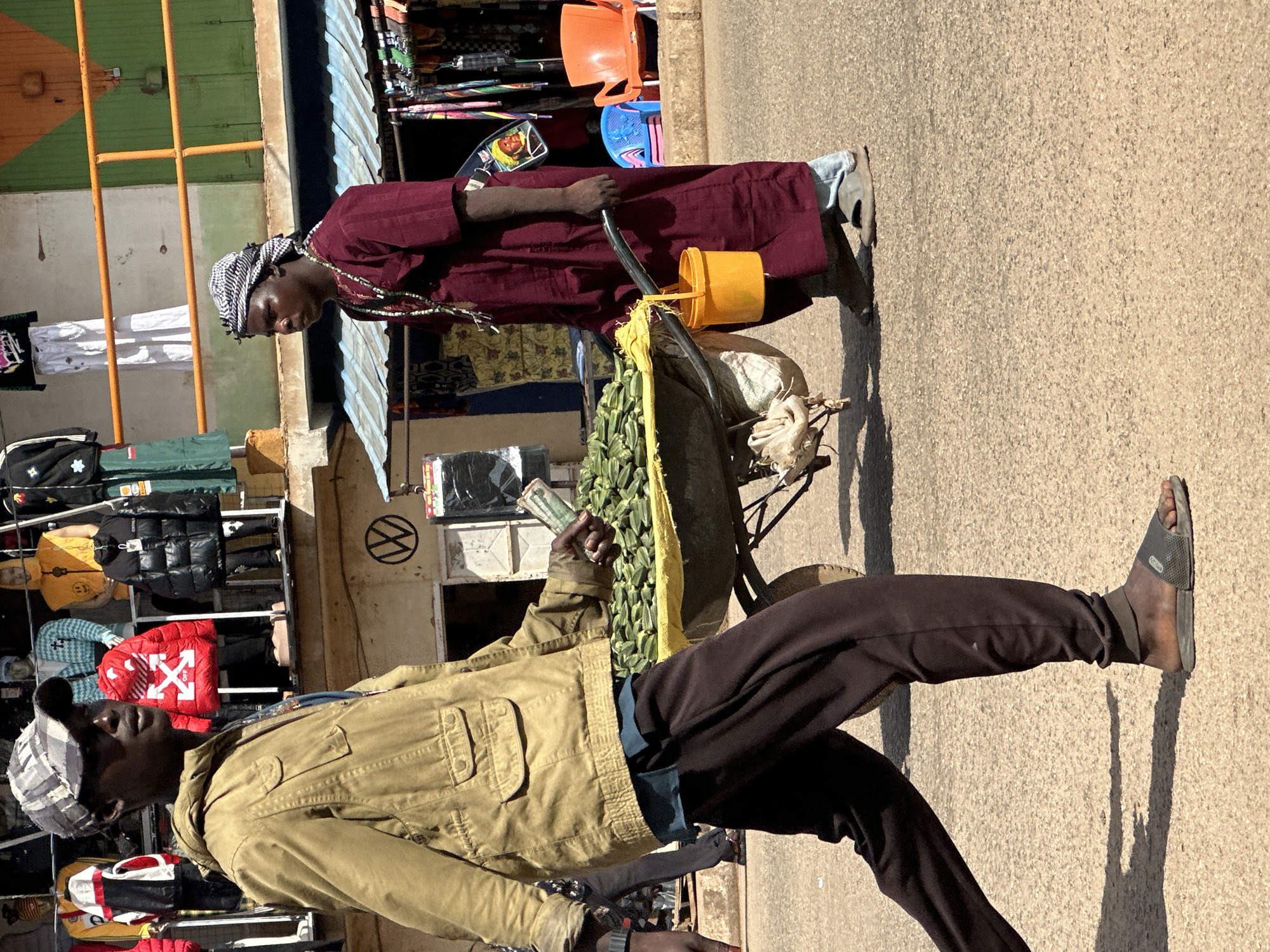In Bukuru, Nigeria, a man wearing a camouflage baseball cap and a tan jacket walks past a market stall. He holds what appears to be money in his right hand. To his right, another man in a maroon robe and headscarf pushes a wheelbarrow filled with green okra. A yellow bucket is attached to the side of the wheelbarrow. The background features market stalls with various clothing items displayed and orange and blue chairs. The ground is paved with asphalt.