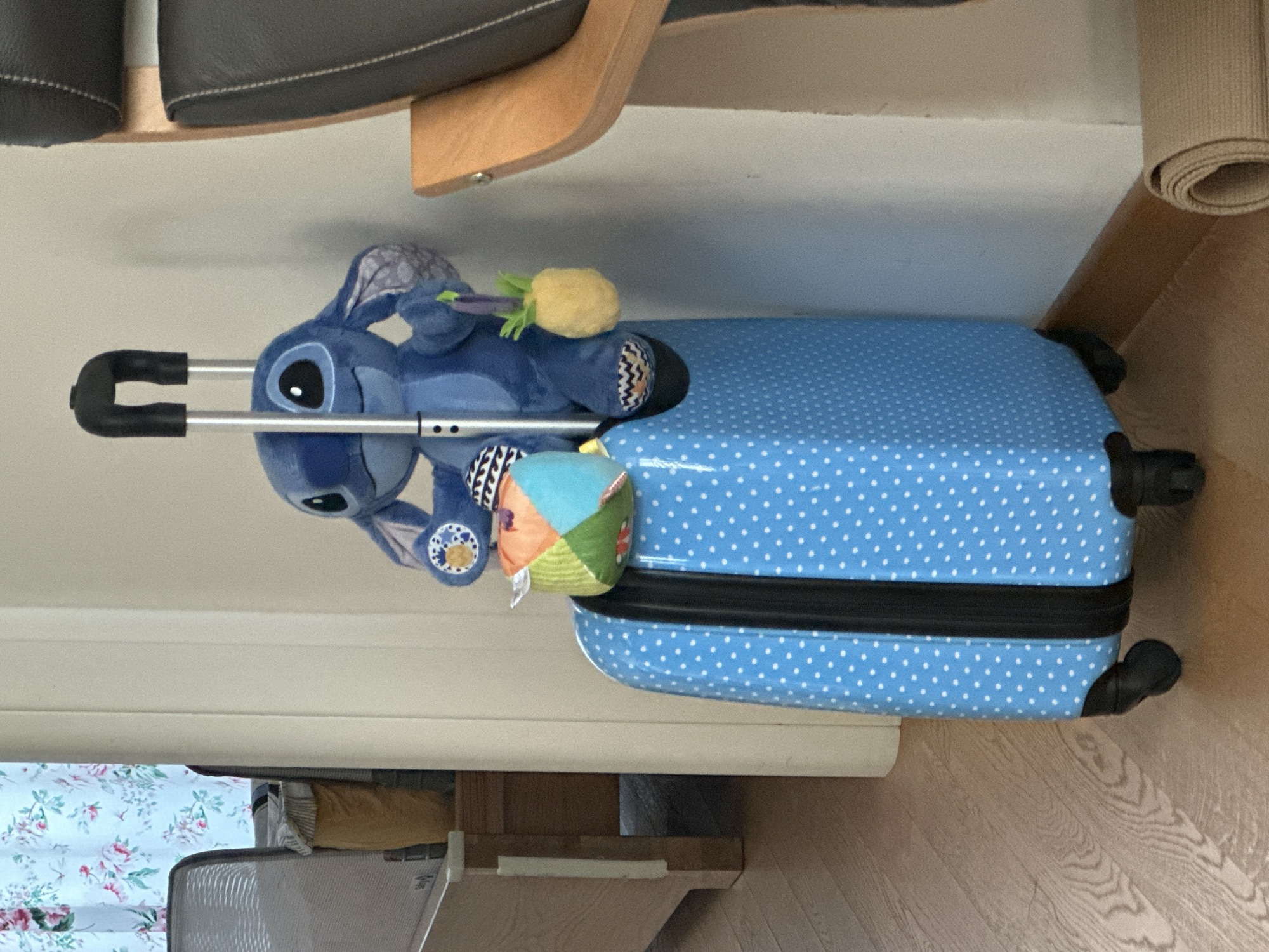 A stuffed Stitch toy with a pineapple charm and a colorful ball is sitting on top of a blue suitcase with white polka dots. The suitcase has a black zipper and black wheels. The scene appears to be indoors, with a wooden floor and a wall in the background. There is a chair with dark upholstery and a wooden frame to the right of the suitcase. To the left, a doorway with floral curtains is visible, along with part of a wooden bed frame. The overall lighting suggests an indoor environment, possibly a home or hotel room. There are no identifiable people in the image. The image does not provide any specific visual cues that would place it in Trieste, Italy.