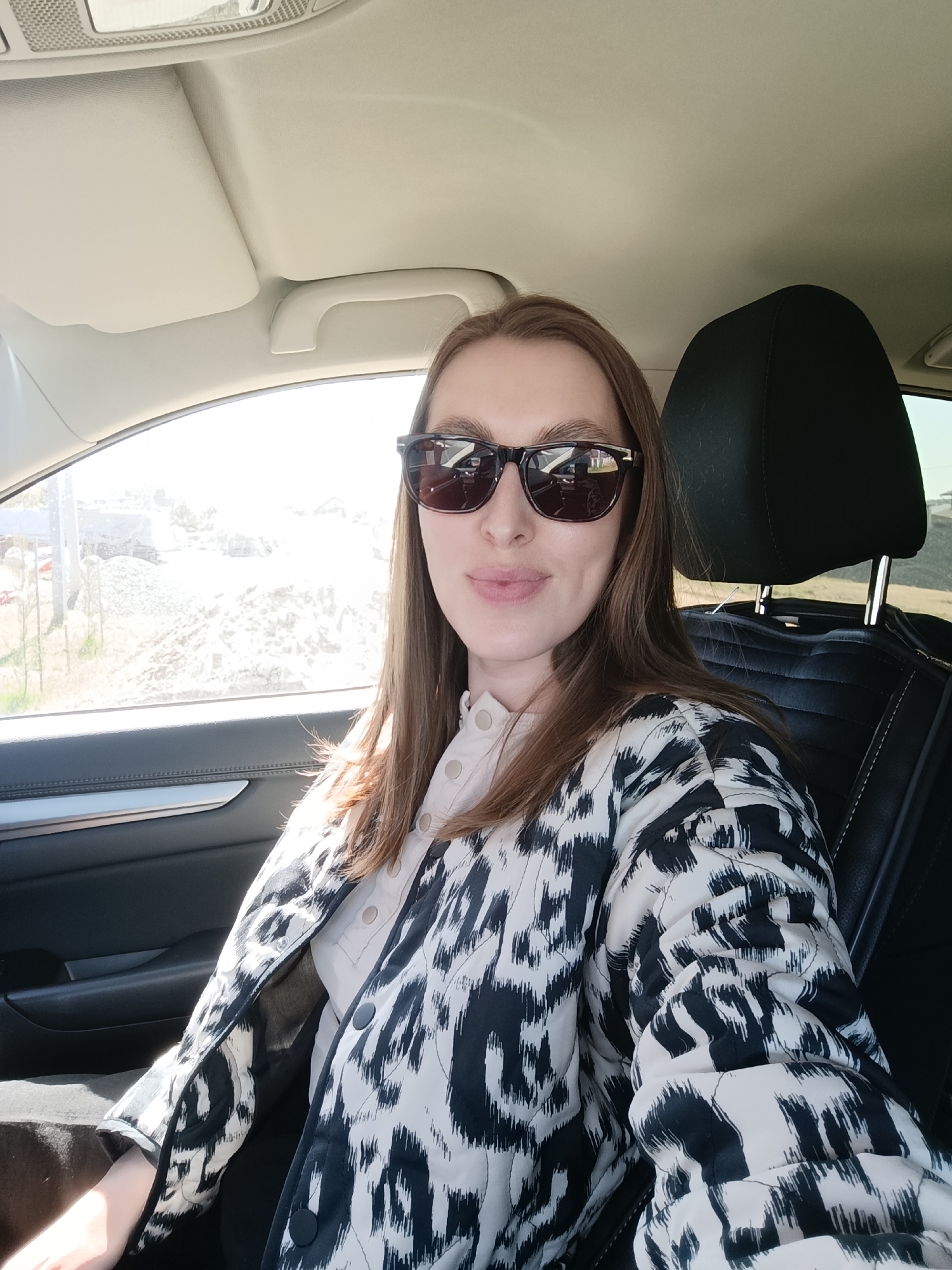 A woman wearing sunglasses and a patterned jacket sits in the driver's seat of a car. Outside the car window, the bright daylight illuminates a construction site with piles of gravel and construction materials. The interior of the car is visible, including the headrest of the driver's seat, the dashboard, and the door panel. Given the location specified in the query, it is inferred that this scene takes place in Chaikovsky, Russia.