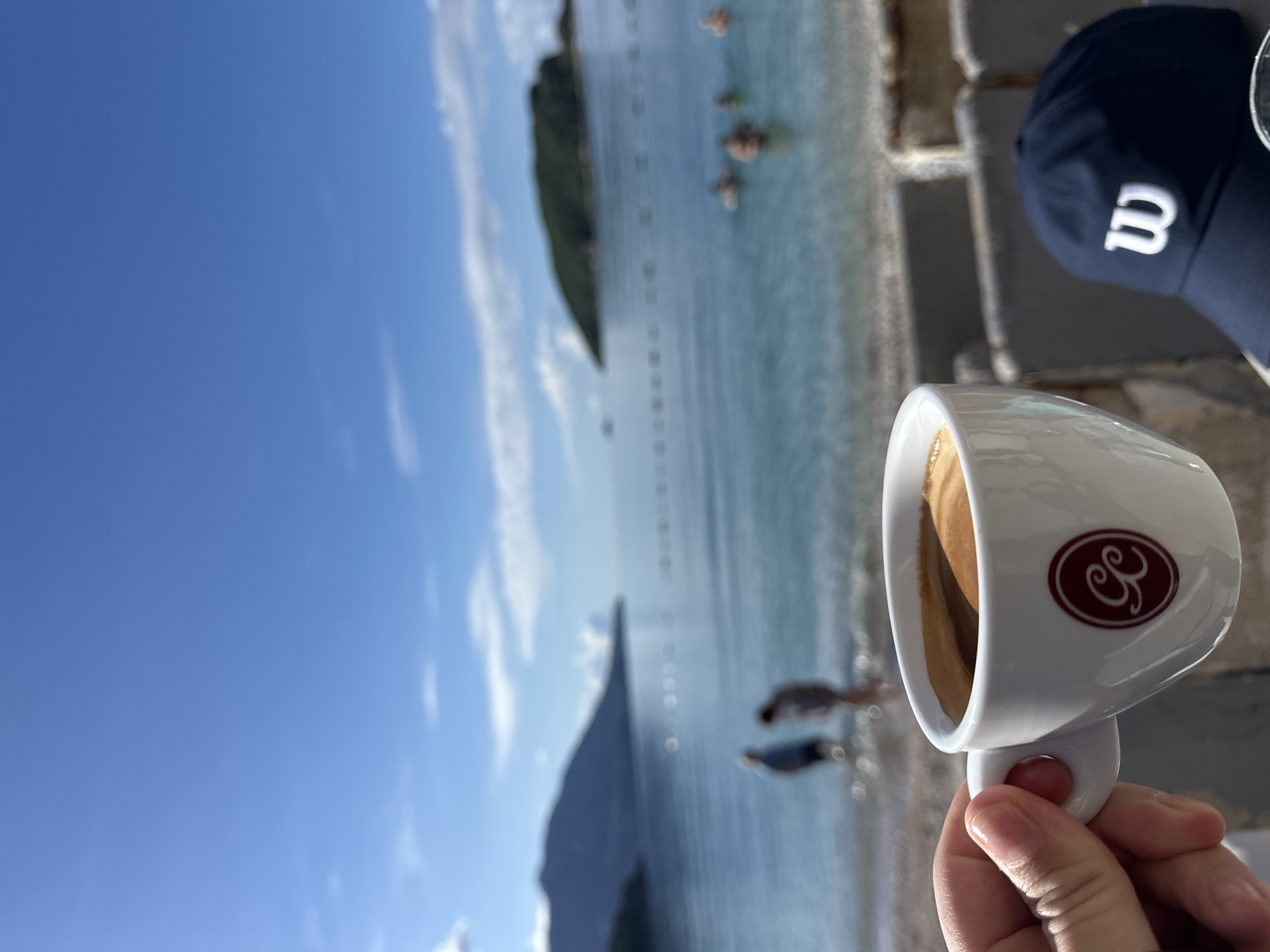 The image shows a person holding a cup of coffee in front of a beach scene. The coffee cup is white with a logo, and the beverage inside appears to be espresso. A hand holds the cup by its handle. The beach background includes clear blue water, a blue sky, a peninsula with vegetation, and indistinct figures of people. A navy blue baseball cap with a white "B" is in the upper right corner. The setting appears to be a cafe or outdoor seating area overlooking the beach. The location is likely near Budva, Montenegro, given the typical landscapes and beach scenery.