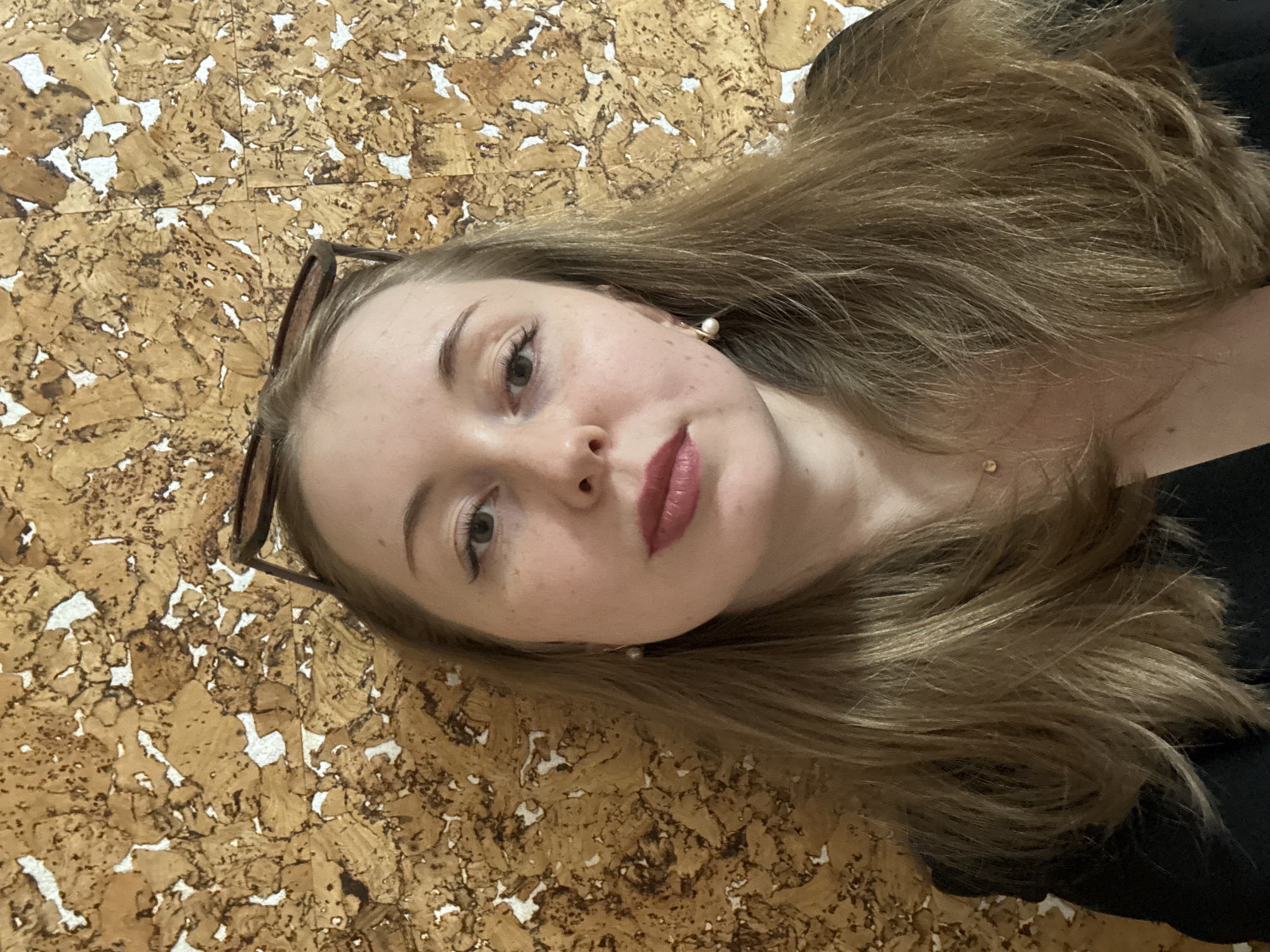 The image is a headshot portrait of a young woman, captured from a slightly high angle and presented rotated 90 degrees counter-clockwise. The woman's head is tilted towards the right side of the frame, and she gazes directly forward. She has fair skin, with visible freckles across her nose and cheeks, and light-colored eyes that appear to be green or hazel. Her lips are full, accentuated with dark pink lipstick. She possesses long, wavy hair in shades of light brown and blonde, flowing behind her and over her left shoulder.
A pair of brown-framed glasses, possibly sunglasses, are resting on top of her head. She wears small, elegant pearl earrings set in gold-colored metal, and a delicate gold chain necklace is faintly visible around her neck. A dark-colored garment, likely a top or dress, is partially visible on her right side. The background is a distinctive textured surface that resembles a cork board or a mosaic of irregularly shaped, light brown and white pieces, creating an abstract pattern. The lighting is soft and even, suggesting an indoor setting or diffused natural light. The woman's expression is neutral and composed. There is no visible text in the image.