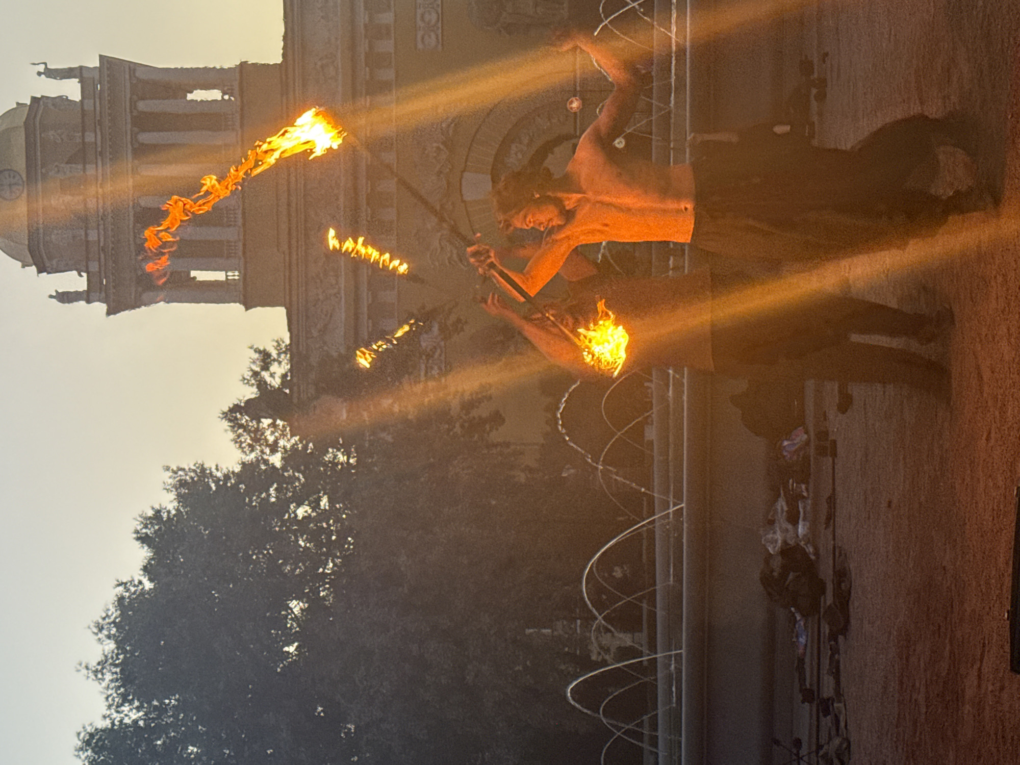 In Saint Petersburg, Russia, two men are fire-spinning in front of a building with a domed roof and classical architecture, possibly at sunset. The building has columns and statues. In the foreground, there is a fountain with arches of water. The ground is covered in dirt or sand. The men are holding flaming staffs, and one is bare-chested, wearing dark pants. The other man is partially obscured and also appears to be holding a flaming staff. There are scattered objects on the ground behind the men, including what appears to be bags and clothing. The scene is illuminated by the flames and the setting sun, creating strong light rays and a warm, orange glow.