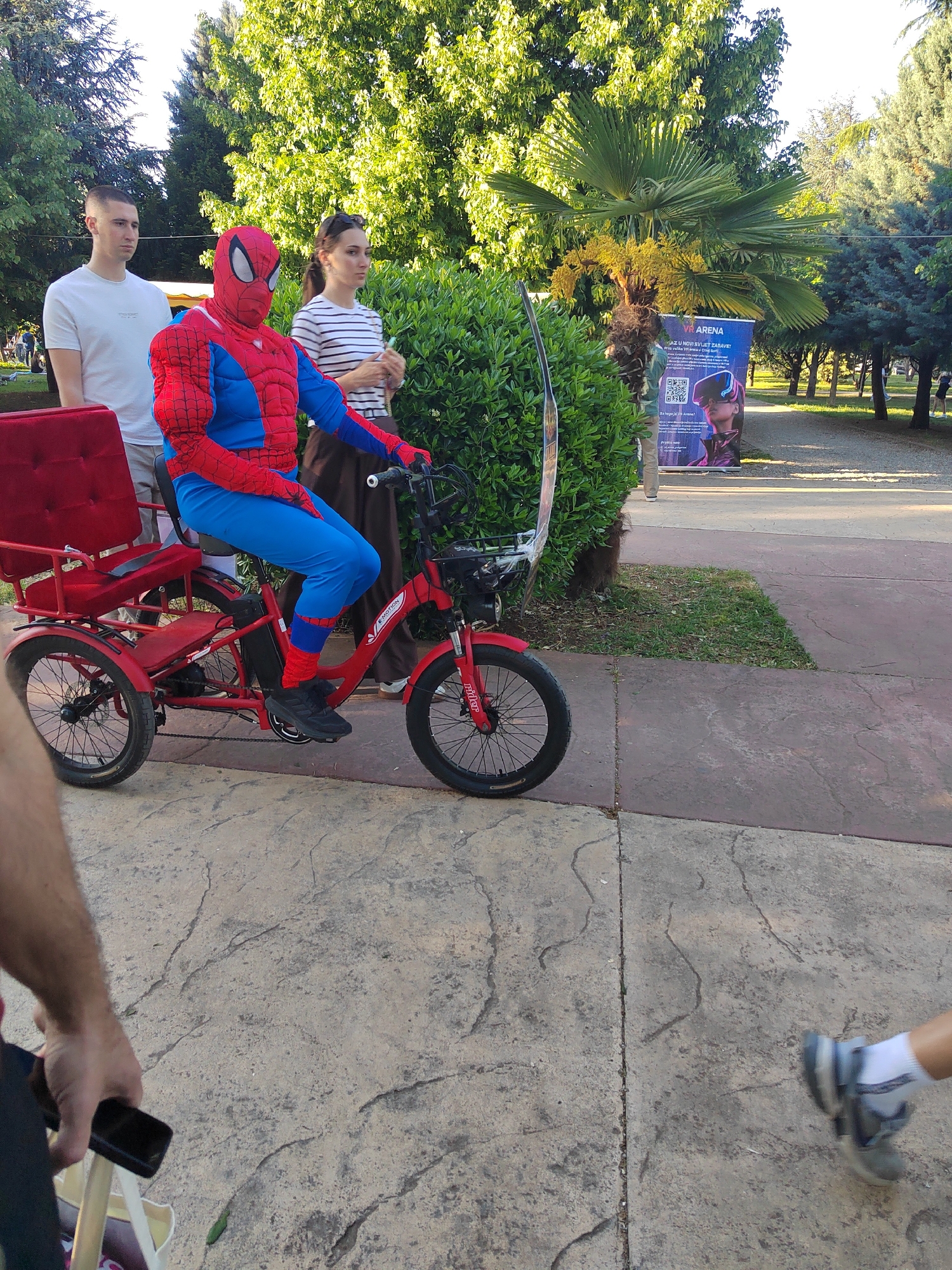 A person dressed as Spider-Man is sitting on a red three-wheeled bicycle on a stone-paved walkway. To his left, a young man in a white t-shirt stands beside the bicycle. To Spider-Man's right, a woman in a striped shirt stands near a bush. The setting appears to be a park or public area in Podgorica, Montenegro, as indicated by the "VR Arena" banner. Additional pedestrians are visible on the path in the background. Another individual with a mobile phone is in the foreground to the left.