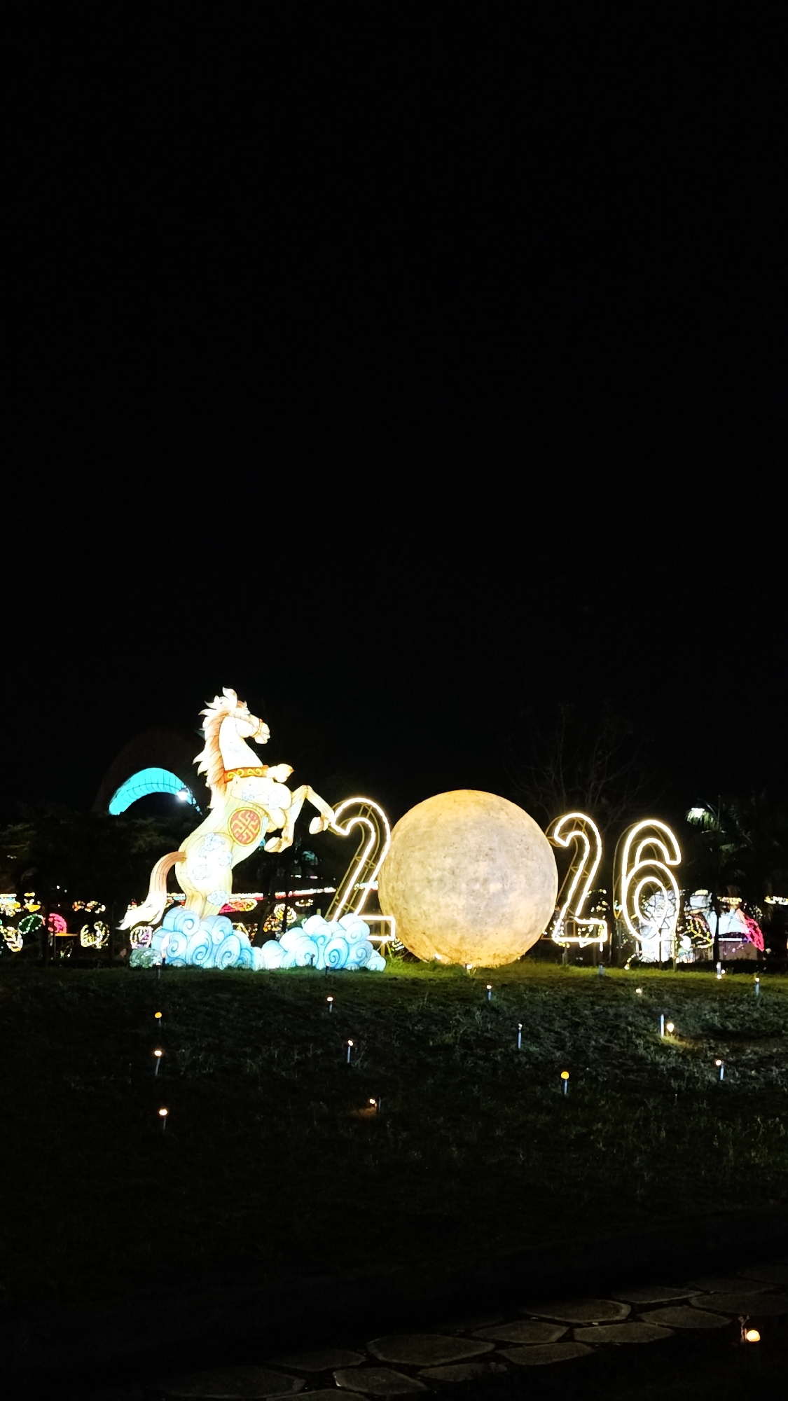At night in Hội An Ward, Vietnam, an outdoor display on a grassy lawn features illuminated figures and numbers. A prancing white horse with ornate designs and a cloud-like base stands to the left of a large spherical light. To the right of the sphere are the numbers "2" and "6" outlined in light. Small lights are sparsely placed in the grass. A blue structure is in the distant background.