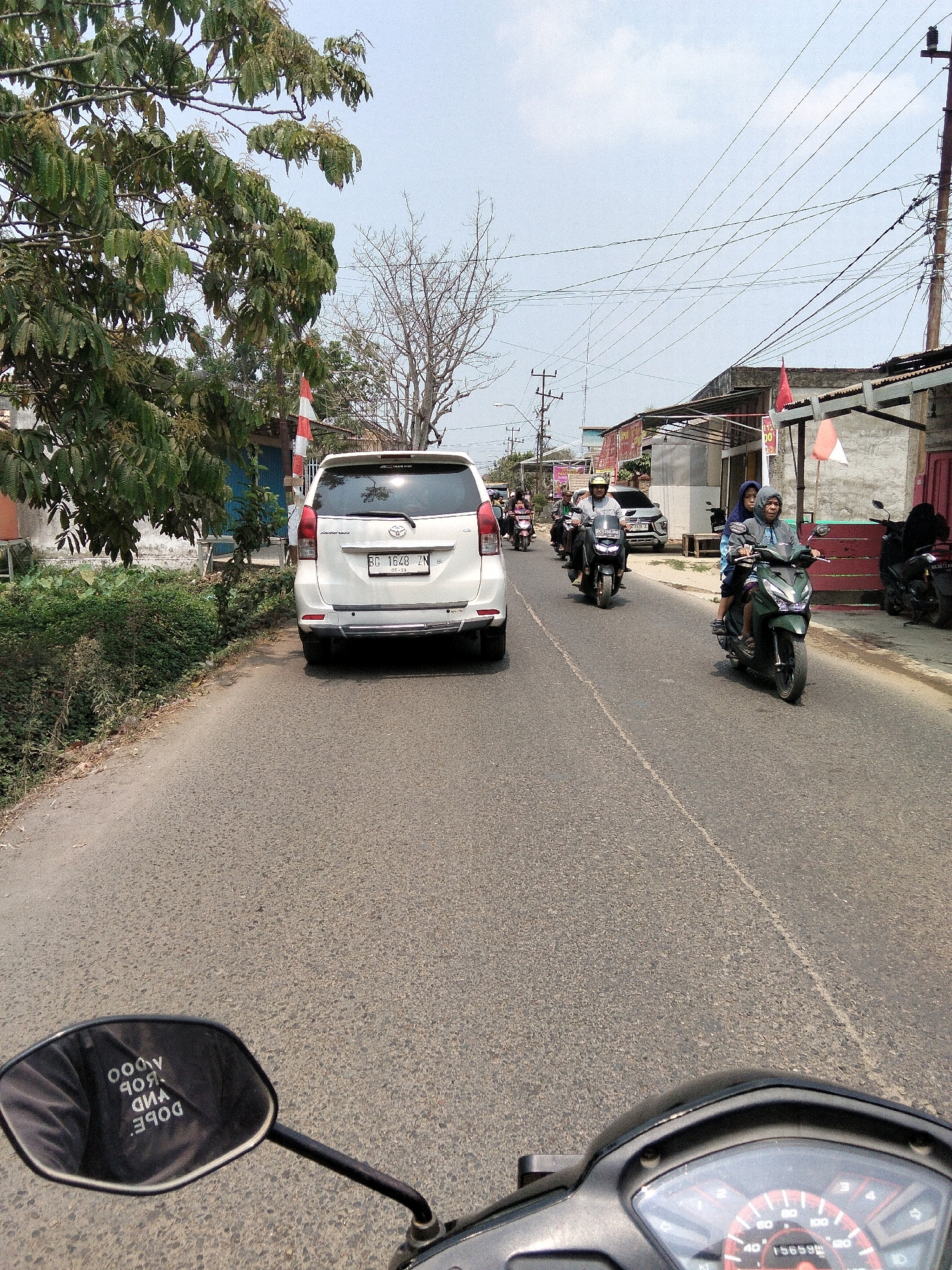 The image depicts a street scene in Sungai Gerong, Indonesia. The perspective is from the viewpoint of someone on a motorcycle, as indicated by the motorcycle's mirror and dashboard visible in the foreground. The mirror reflects text that reads "DOPE. VIBD ROBY ADOO". A white SUV is parked on the side of the road, with a license plate reading "BG 1648 ZM". Several motorcycles are also present, with riders and passengers. One green motorcycle with two people is in motion, as is a black motorcycle with a rider further ahead. The road is paved, and the environment appears to be a mix of residential and commercial buildings, with trees and power lines visible. The sky is clear and blue.