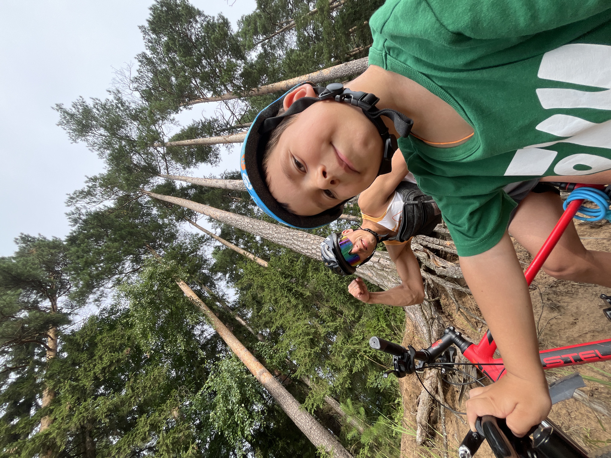 In the city of Chaikovsky, Russia, two individuals are pictured amidst a dense forest with tall pine trees. The foreground features a young boy wearing a blue helmet and a green t-shirt with white lettering, holding onto the handlebars of a red bicycle. To his left and slightly behind, a man with a helmet and sunglasses is flexing his bicep. He is wearing a white tank top and a black vest with a fanny pack. Both individuals appear to be on a dirt path, with exposed tree roots visible. The sky is overcast.
