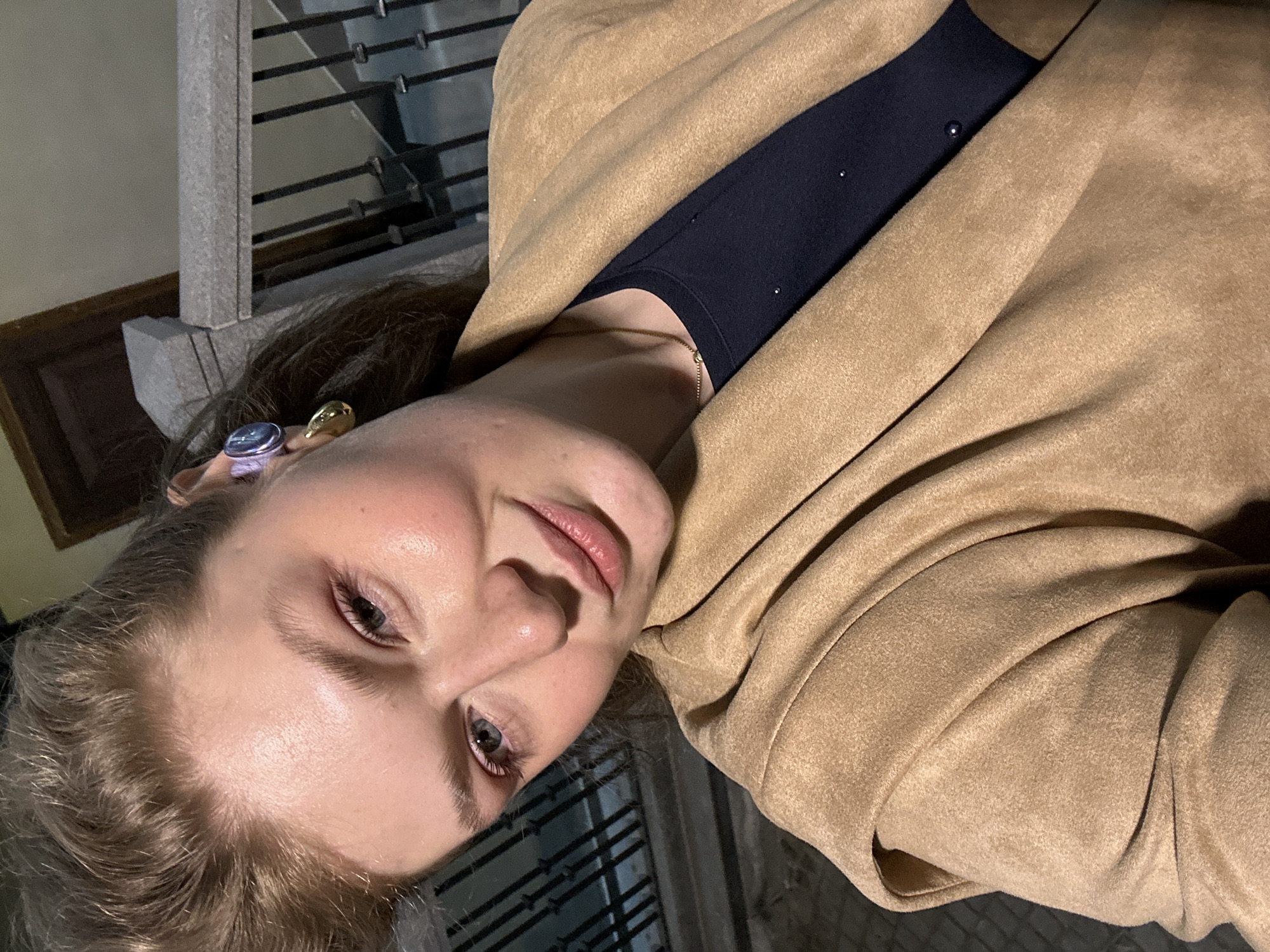 A person is depicted in a close-up, low-angle shot wearing a tan-colored suede or brushed fabric coat or jacket. Underneath the coat, they are wearing a black top. The person has fair skin and light-colored hair and is wearing two distinctly colored earrings, one purple and one gold. A thin gold necklace is visible at their neck. The background shows a metal railing and what appears to be a building facade, possibly indicating an outdoor location. The image is taken at night or in low light. There is no verifiable information present to determine if the image was taken in Kazan, Russia.
