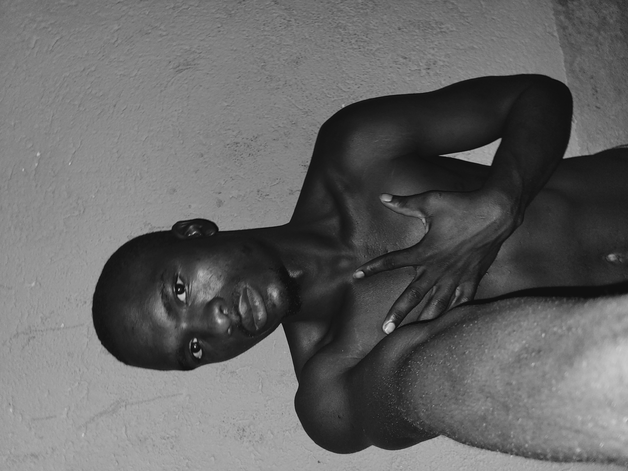 A grayscale, close-up, waist-up shot captures a young Black man, shirtless, in what appears to be an indoor setting. The man's head is slightly tilted, and he is looking directly at the camera with a neutral expression. His right arm is raised and bent, with his hand placed on his chest, fingers spread in a V-shape. His left arm is extended forward and out of frame, the bicep appears to be glistening with sweat. The background is a textured wall, dimly lit and nondescript. There is no identifiable signage or landmarks to confirm a specific location in Bukuru, Nigeria.