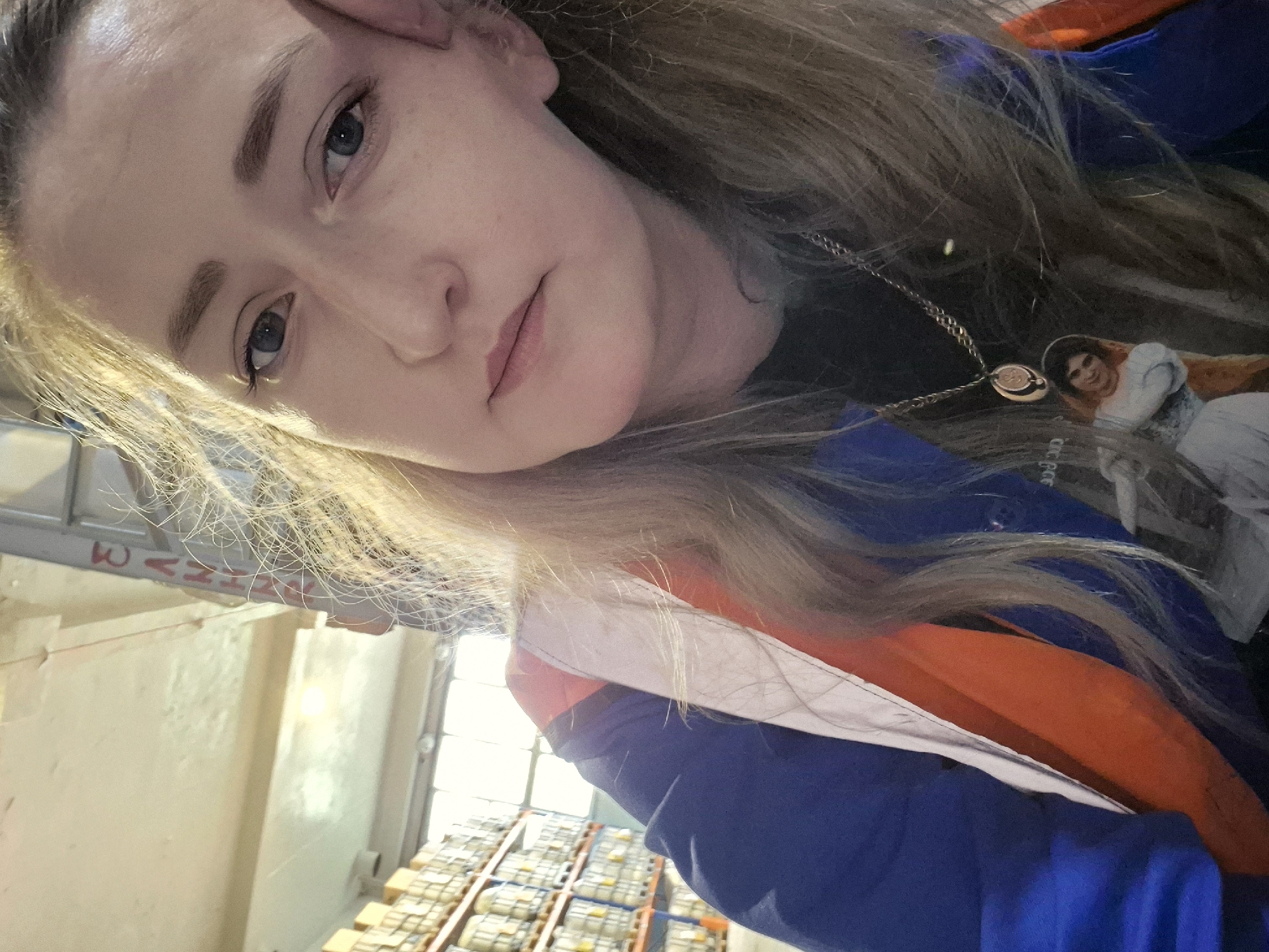 A young Caucasian woman with blonde hair and blue eyes is shown in a selfie. She is wearing a blue jacket with orange and white accents, and a silver necklace with a pendant. Her right hand is resting on her cheek. In the background, shelves stacked with yellow boxes are visible, suggesting an industrial or warehouse setting. There are also some faint red markings on a vertical structure in the background. The lighting suggests it is daytime. The scene is likely located in a warehouse in Chaikovsky, Russia, given the mention of that location.