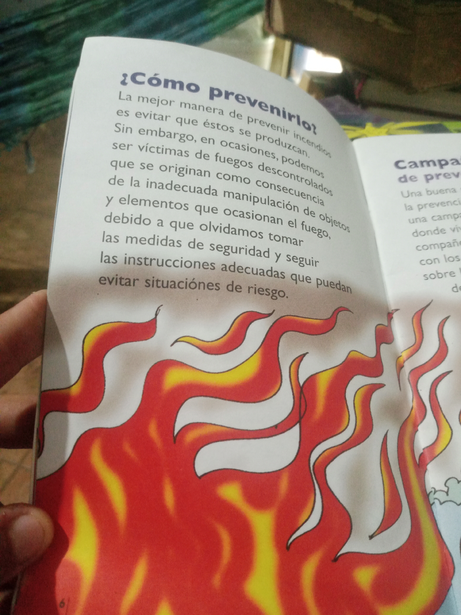 The image shows two pages of an open book, featuring text and illustrations. The left page has a large heading in Spanish, "¿Cómo prevenirlo?" (How to prevent it?), followed by several paragraphs of text discussing fire prevention. Below the text, there is a stylized illustration of red and yellow flames. The right page features a heading in Spanish, "Campan" (likely part of "Campañas" meaning campaigns), and some text beneath it. This page also has an illustration of stylized blue and white clouds, suggesting a contrast with the fire on the left. The book is open, and a hand is visible on the far left of the image, holding the book open. The background appears to be an indoor setting with some blurred objects, possibly furniture or other books. There is no visible indication of the city of Santa Ana, Venezuela, in the image. The scene is a close-up of an open book with illustrative content related to fire safety.