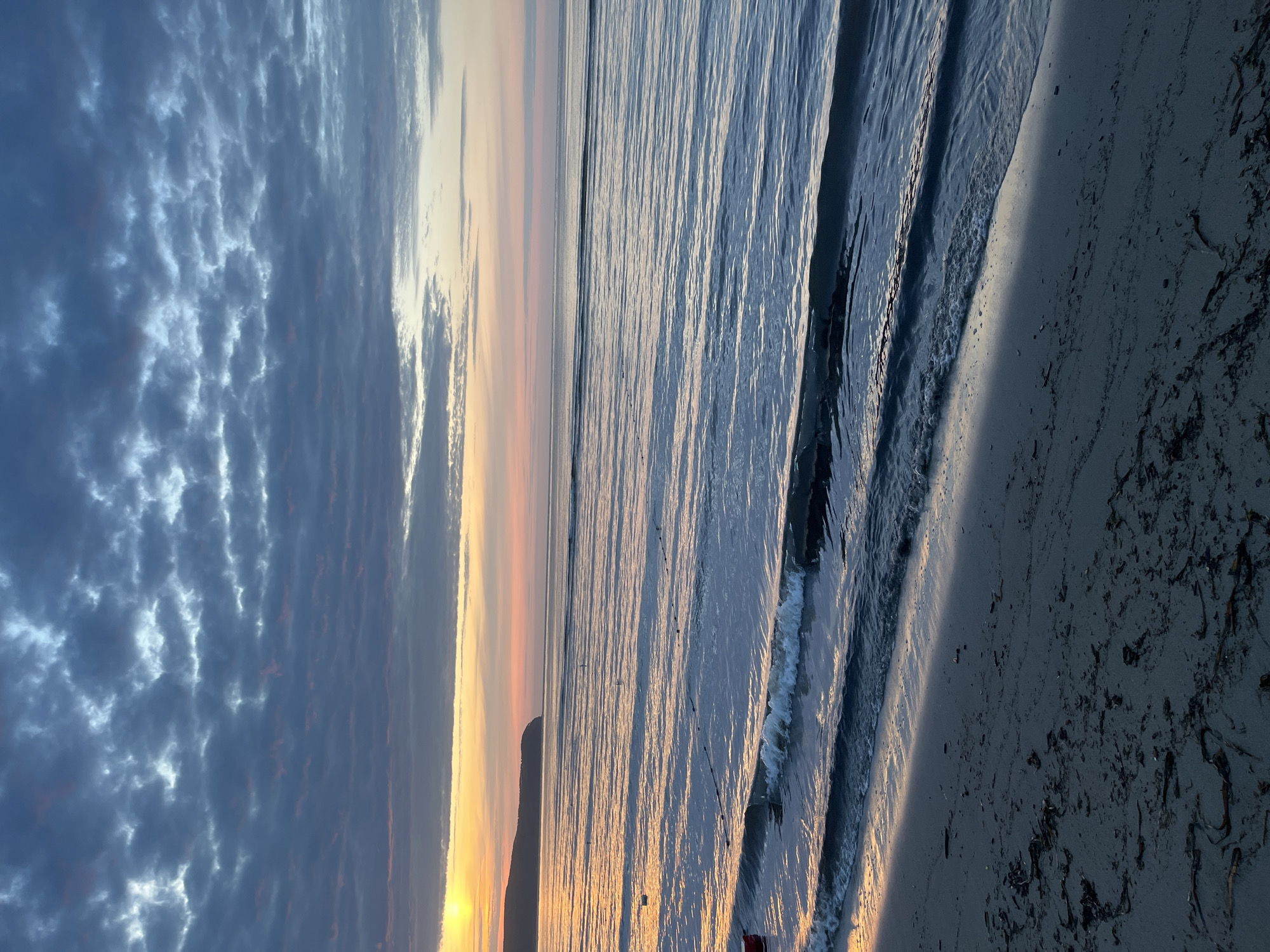 The image depicts a beach scene at sunset. The sky is filled with dramatic clouds, tinged with hues of orange and pink, suggesting the sun is low on the horizon. The ocean is calm, with gentle waves lapping at the shore. The wet sand of the beach reflects the warm light of the sunset, creating a shimmering effect. Scattered seaweed and debris are visible on the darker, drier sand further up the beach. In the distance, a silhouette of land or a hill can be seen against the vibrant sky. The overall mood is serene and peaceful, characteristic of a sunset on a beach. The image is oriented vertically, as if taken with a phone held in portrait mode.