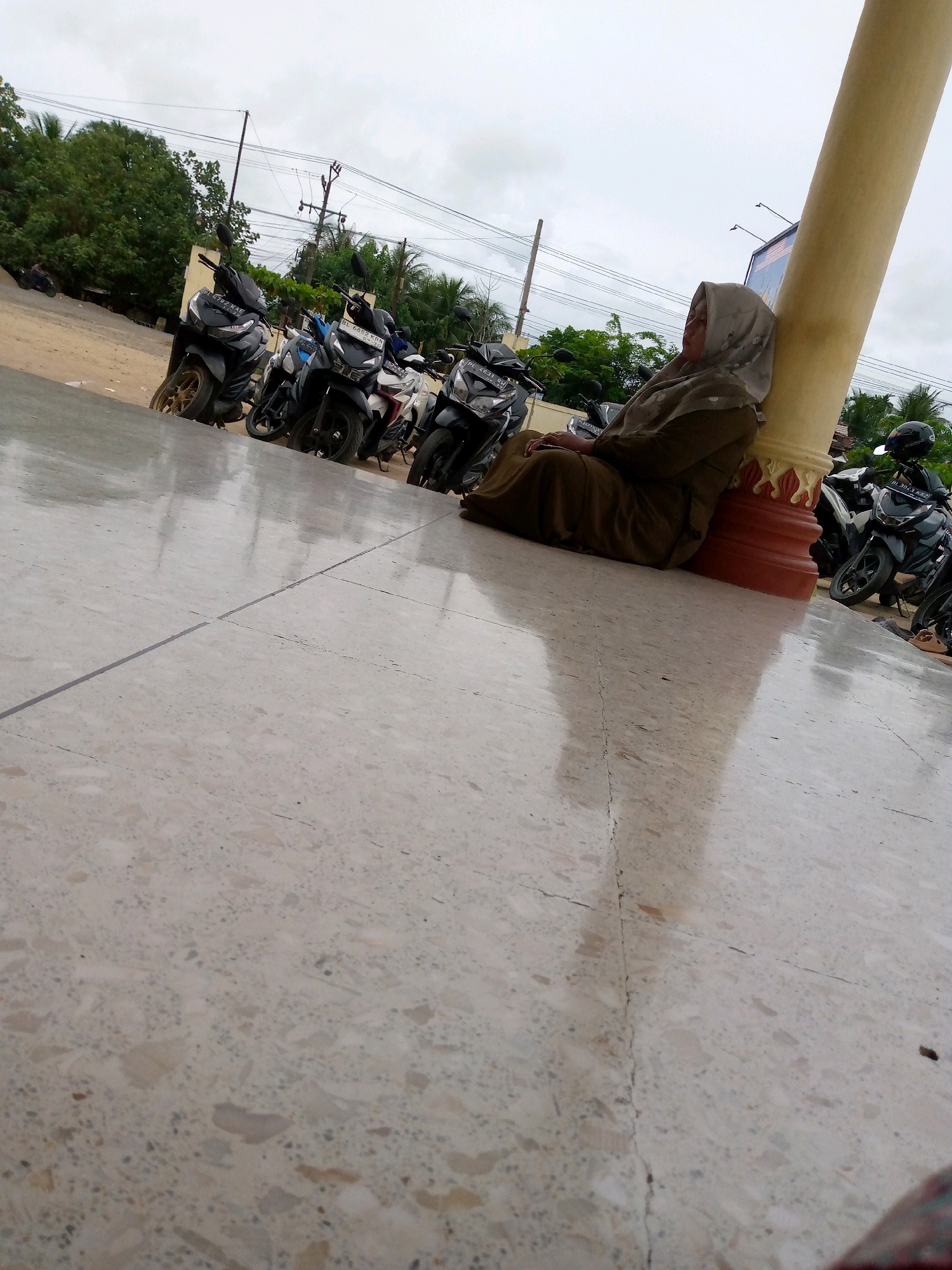 In Keudeteupinpunti, Indonesia, a person is sitting on a patterned tile floor, leaning against a large, yellow, cylindrical pillar. The individual is wearing a light brown hijab and a matching robe, covering their entire body except for their face. The person appears to be resting or waiting, with their eyes closed. Behind them, a row of parked motorcycles is visible, suggesting an outdoor public area, possibly a parking lot or a communal space. The sky is overcast, indicating a cloudy day. The ground is reflective, possibly due to recent rain or a polished surface. The overall atmosphere is tranquil and still.