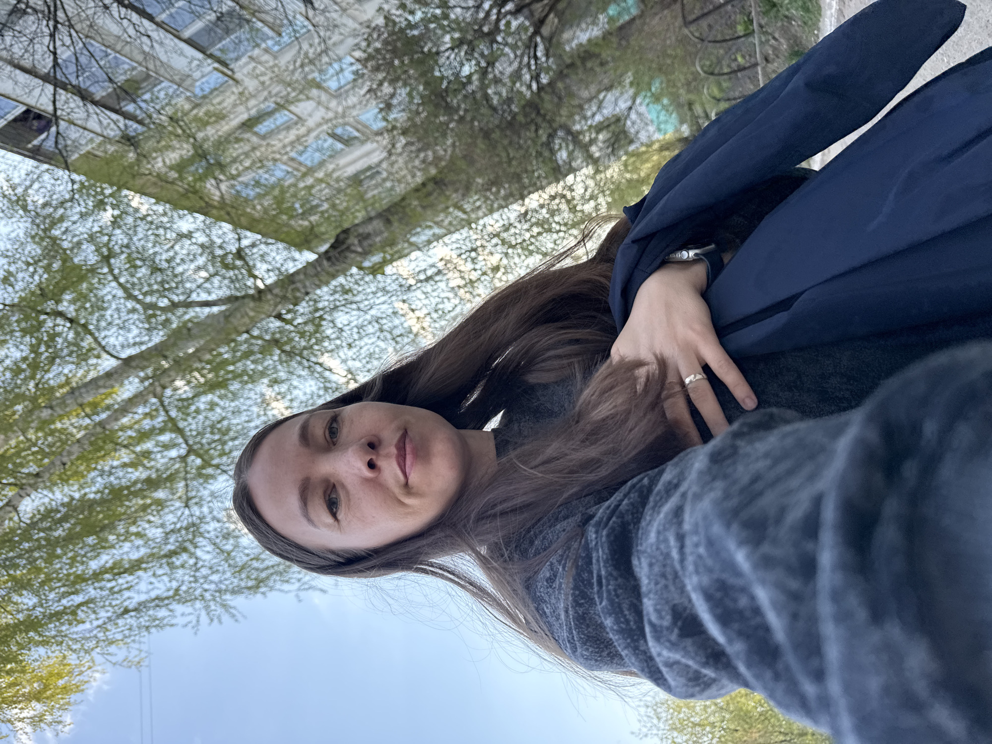 A photograph taken in Chaikovsky, Russia, shows a woman from a low angle perspective. The woman has long brown hair and is wearing a gray sweater. She also has a blue jacket draped over her arm. She has a ring on her finger. In the background is a building, trees with new leaves, and a clear blue sky.