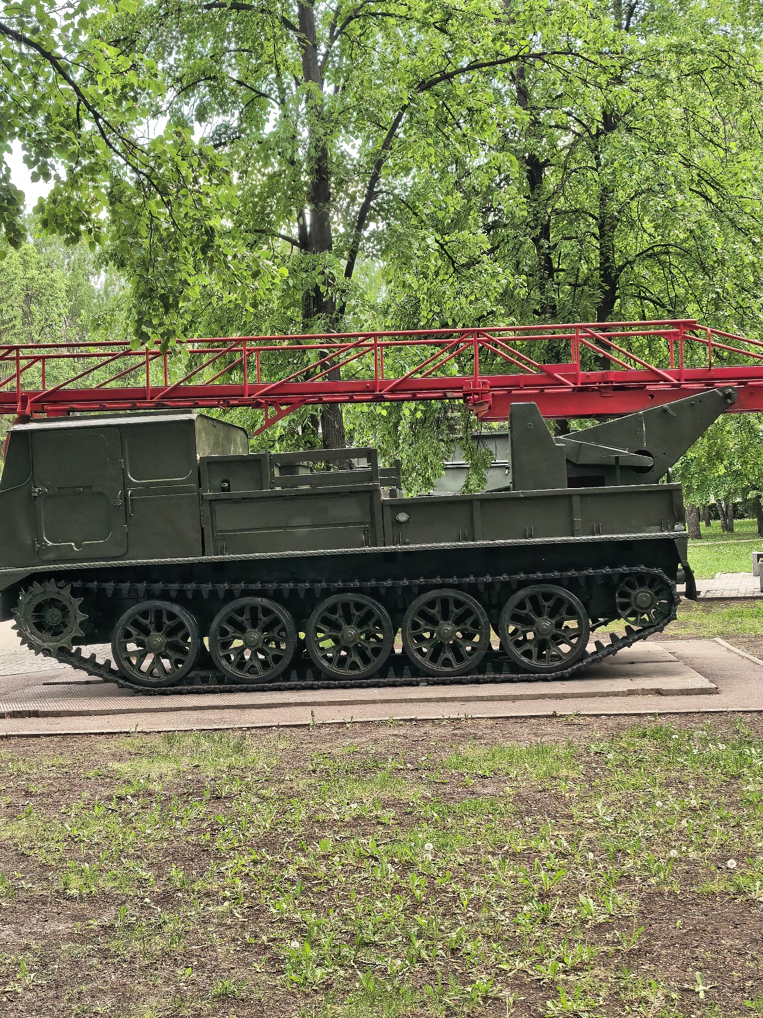 The image depicts a preserved military vehicle, specifically an artillery tractor with a red folding bridge mounted on its chassis. The vehicle is olive green, with prominent tracks and multiple road wheels. It rests on a low concrete platform surrounded by a grassy area with scattered vegetation and bare patches of soil.
In the background, lush green trees form a canopy, suggesting a park or green space. The location is a public display, likely a memorial or exhibit. While the city name is provided, no identifying markers are present in the image itself. The scene appears static and unoccupied, with no people or moving elements visible.