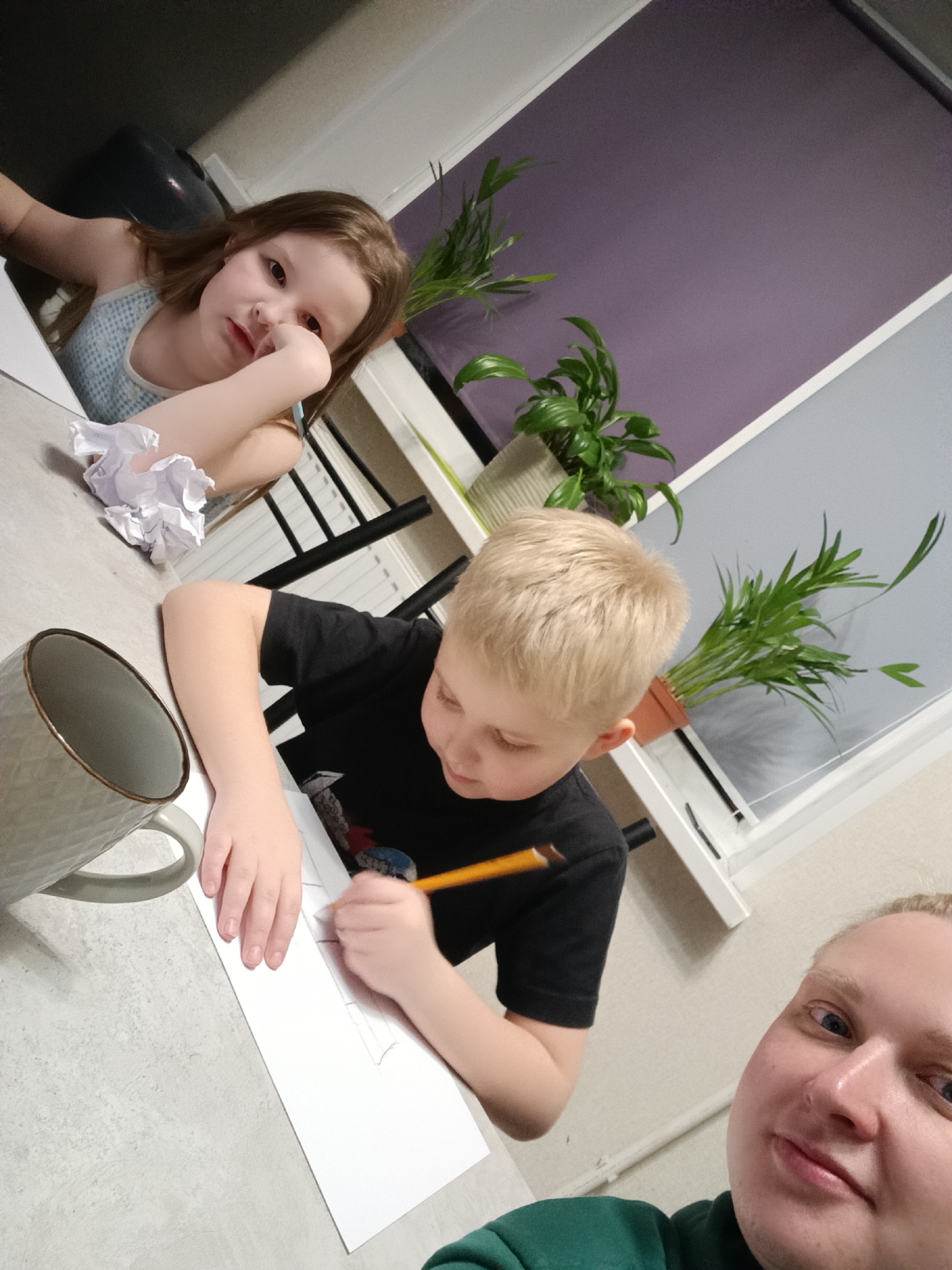 Here's a forensic description of the image:
**Scene:** A tabletop setting. The lighting is artificial.
**People:**
* A young girl with long brown hair is seated, appearing bored or tired, with her head resting on her hand.
* A young boy with blond hair is drawing with a pencil on a sheet of white paper.
* Part of a person's face (likely an adult male) is visible in the foreground, suggesting they are taking the picture. They are wearing a green top.
**Objects:**
* A large mug is present on the table.
* Crumpled pieces of paper are near the girl.
* Several potted plants are visible near a window.
**Actions:**
* The boy is drawing.
**Environment:**
* A window with white frames and what appears to be closed blinds is visible in the background.
There is no verifiable information that the photo was taken in Chaikovsky, Russia.