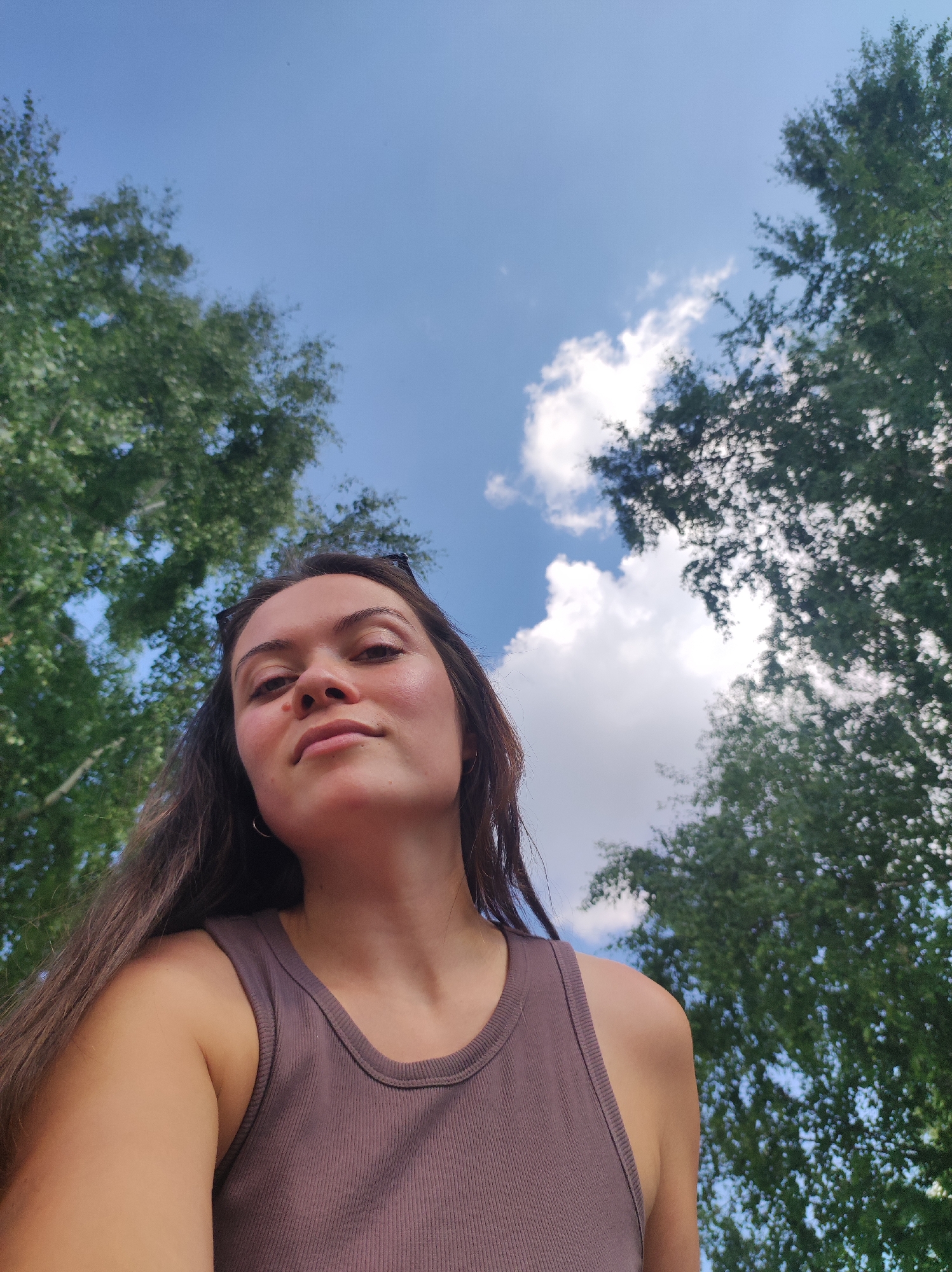A low-angle, medium shot captures a young woman from the chest up, looking directly at the camera. She has long, dark brown hair, and her face is illuminated by natural light, highlighting her features. She is wearing a brown ribbed tank top and a silver hoop earring is visible in her left ear. Her pose is casual, with her head slightly tilted.
The background consists of a bright blue sky with scattered white clouds and the dense green foliage of trees. The trees frame the top and sides of the image, suggesting an outdoor setting, possibly a park or a natural area. The overall impression is a candid shot taken on a sunny day. The image does not contain any specific visual cues to determine its location within the city of Марковский, Russia.