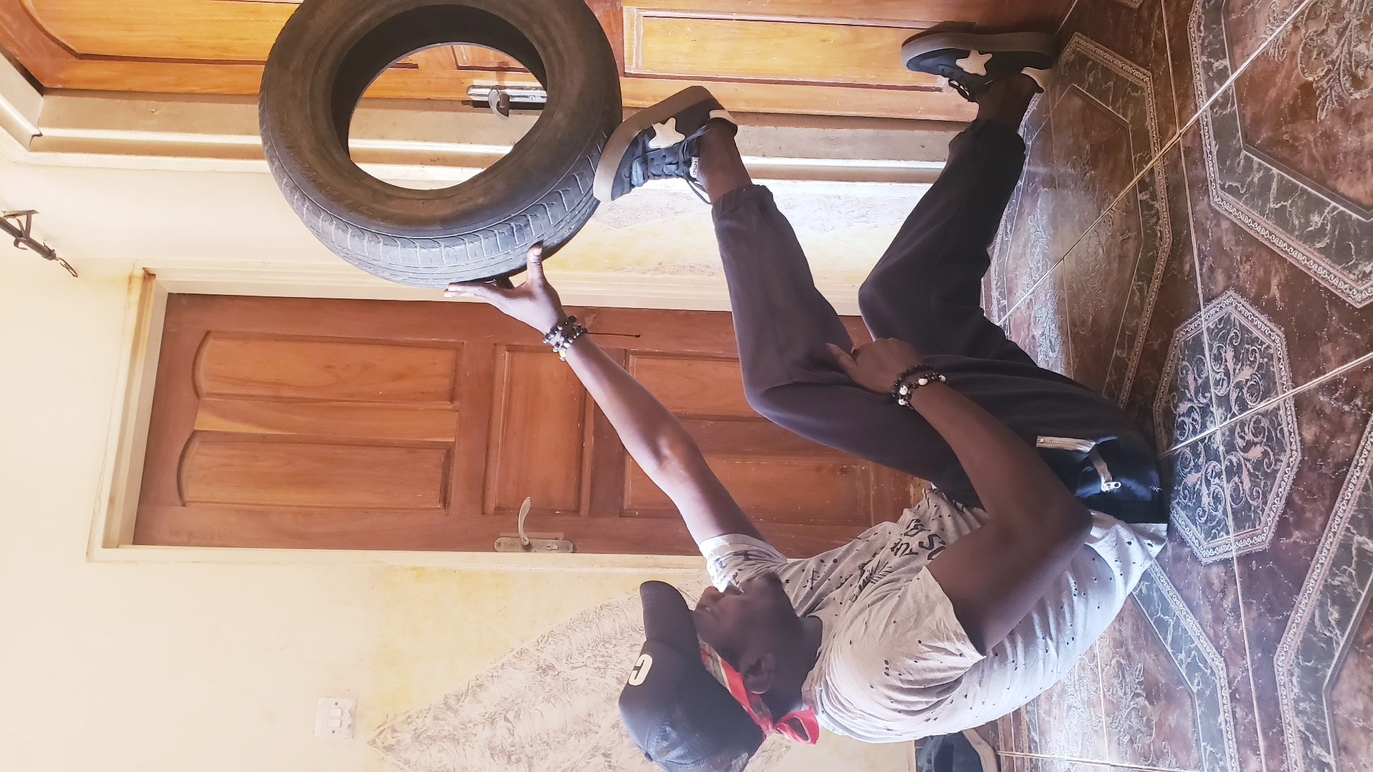 An indoor, full-length shot captures a person balancing a car tire. The image is presented rotated 90 degrees counter-clockwise. When viewed correctly, a young man stands on a patterned, brown-and-black tiled floor, holding a car tire above his head with his right hand. He is dressed in a light-colored t-shirt with a dark pattern, dark trousers, and black sneakers featuring a prominent white star design. A black baseball cap is worn over his head, with a red bandana visible underneath, and he has beaded bracelets on both wrists.
His body is slightly angled, with his left leg slightly bent, and he appears to be positioned near a light brown wooden door with carved panels, which is set into a light yellow wall. A white light switch or outlet is visible on the lower section of this wall. The scene is brightly lit, suggesting daytime. The activity demonstrates a moment of balance or playful skill. No discernible text is visible on clothing or objects. The setting is an indoor space in Sabon Tasha, Nigeria.
