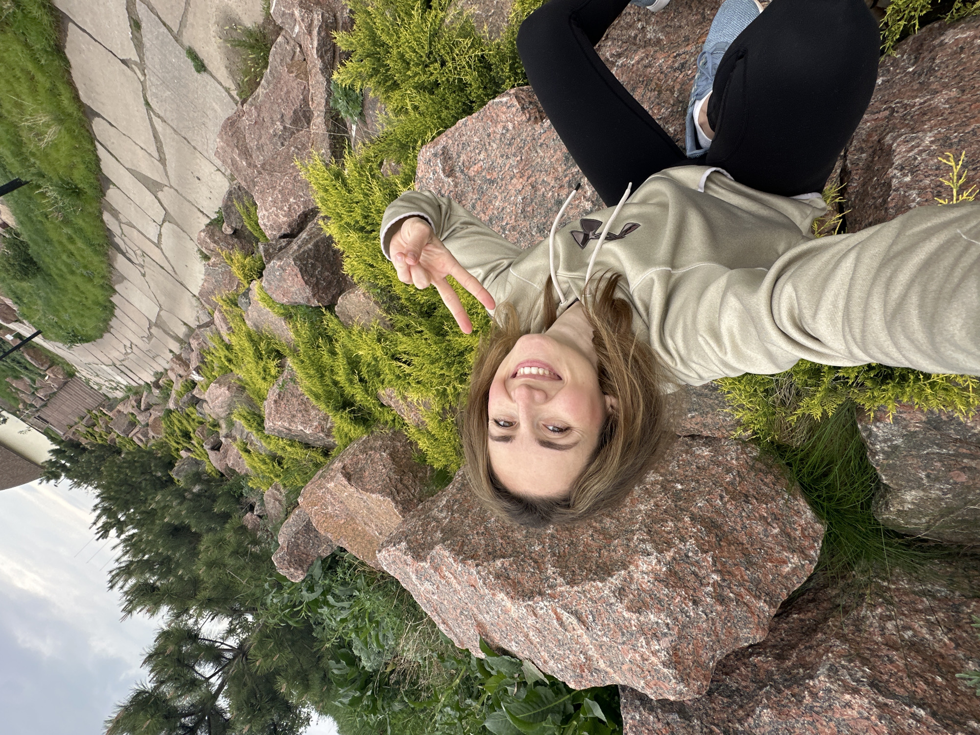 A young woman lies on a large, reddish-brown rock formation in an outdoor setting. She wears black leggings, light blue sneakers, and a light-colored hoodie, and she smiles and makes a peace sign with her right hand. The rocks are interspersed with green shrubbery, appearing to be part of a landscaping design. In the background, there is a stone-paved walkway, green grass, and buildings. The location is Zakharivka, Ukraine.