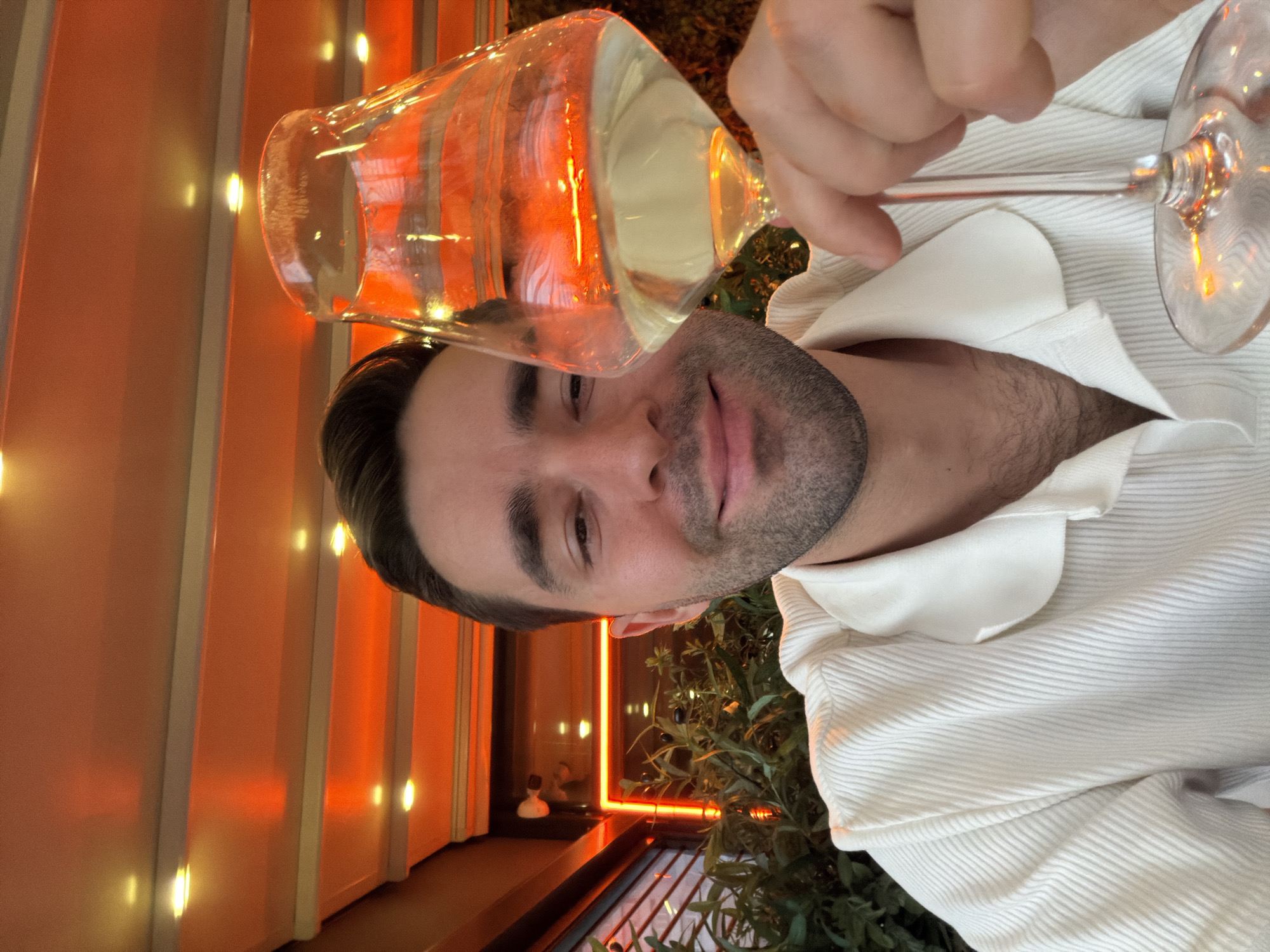 A man is holding a glass of white wine in front of his face. He is wearing a white, textured shirt with a prominent collar. The background includes orange-lit vertical stripes, possibly an interior design feature, and glimpses of greenery, suggesting a restaurant or bar setting. Based on the light and environment, this could be located in the city of Khimki, Russia. The man seems to be posing for the photo.