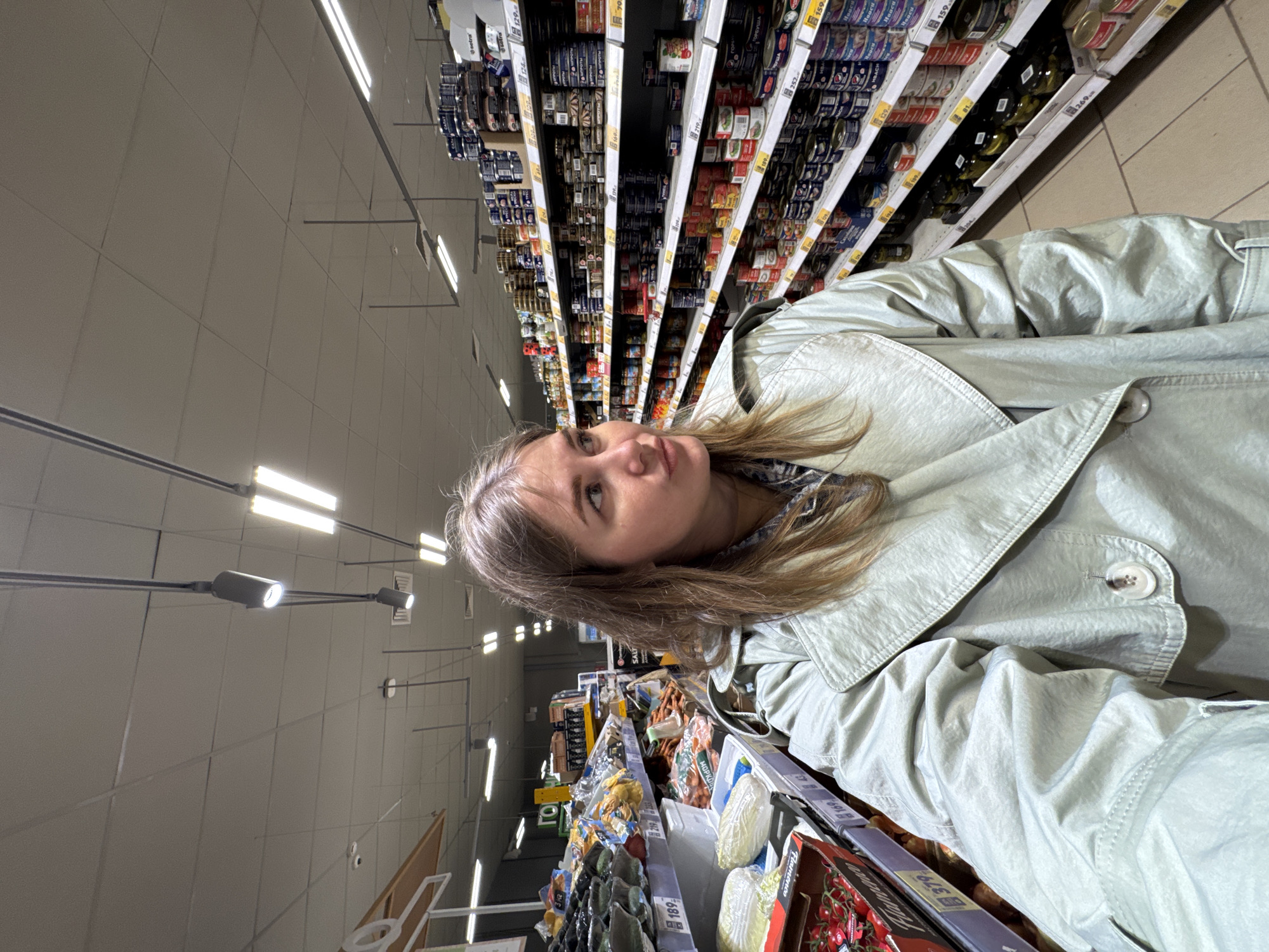 Here is a forensic description of the image:
The image shows a fair-skinned young woman with light brown hair, standing in a grocery store aisle, seemingly taking a selfie or having her picture taken. She is wearing a light-colored, perhaps light green or grey, trench coat.
Behind her are shelves stacked with canned goods. The cans are labeled in what appears to be the Russian language, along with some price tags with prices such as "252" and "159." She appears to be standing next to a display of vegetables, including tomatoes.
A price tag of "379" is clearly visible on the edge of her shopping basket.
The bright fluorescent lighting and overhead structures of a grocery store are seen above the aisles. The location is Горный, Russia, based on the OCR of Russian words.