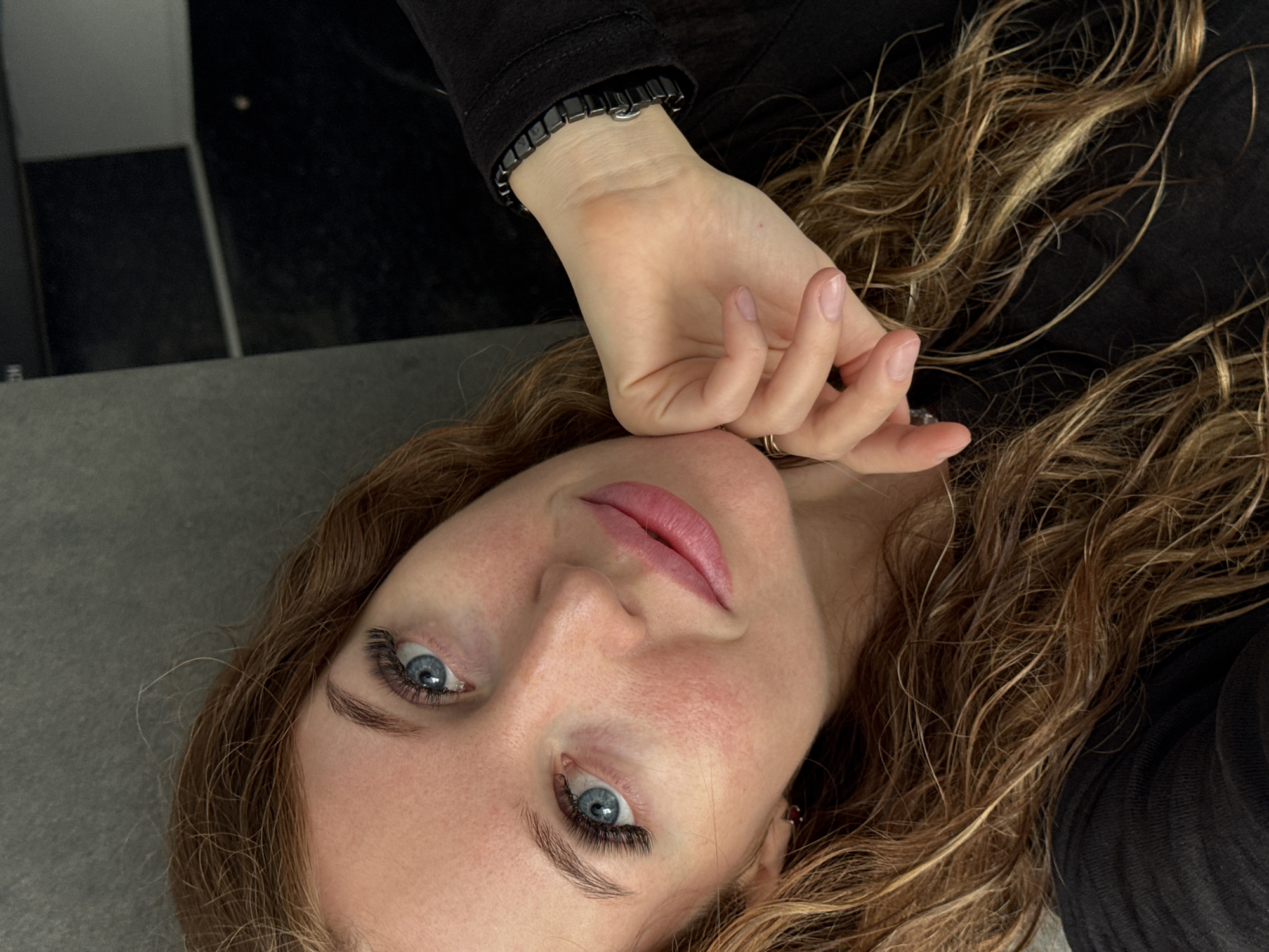 The image shows a close-up, selfie-style shot of a young woman with long, wavy, auburn hair. She has striking blue eyes with prominent false eyelashes, fair skin with visible pores and some redness on her cheeks, and full, pink lips. Her right hand is resting on her cheek, and she is wearing a black long-sleeved top and a black bracelet on her wrist. The background appears to be a dark, textured surface, possibly a piece of furniture or a wall, with some lighter grey areas. There are no discernible features in the image that would indicate a specific location in the city of Chaikovsky, Russia.