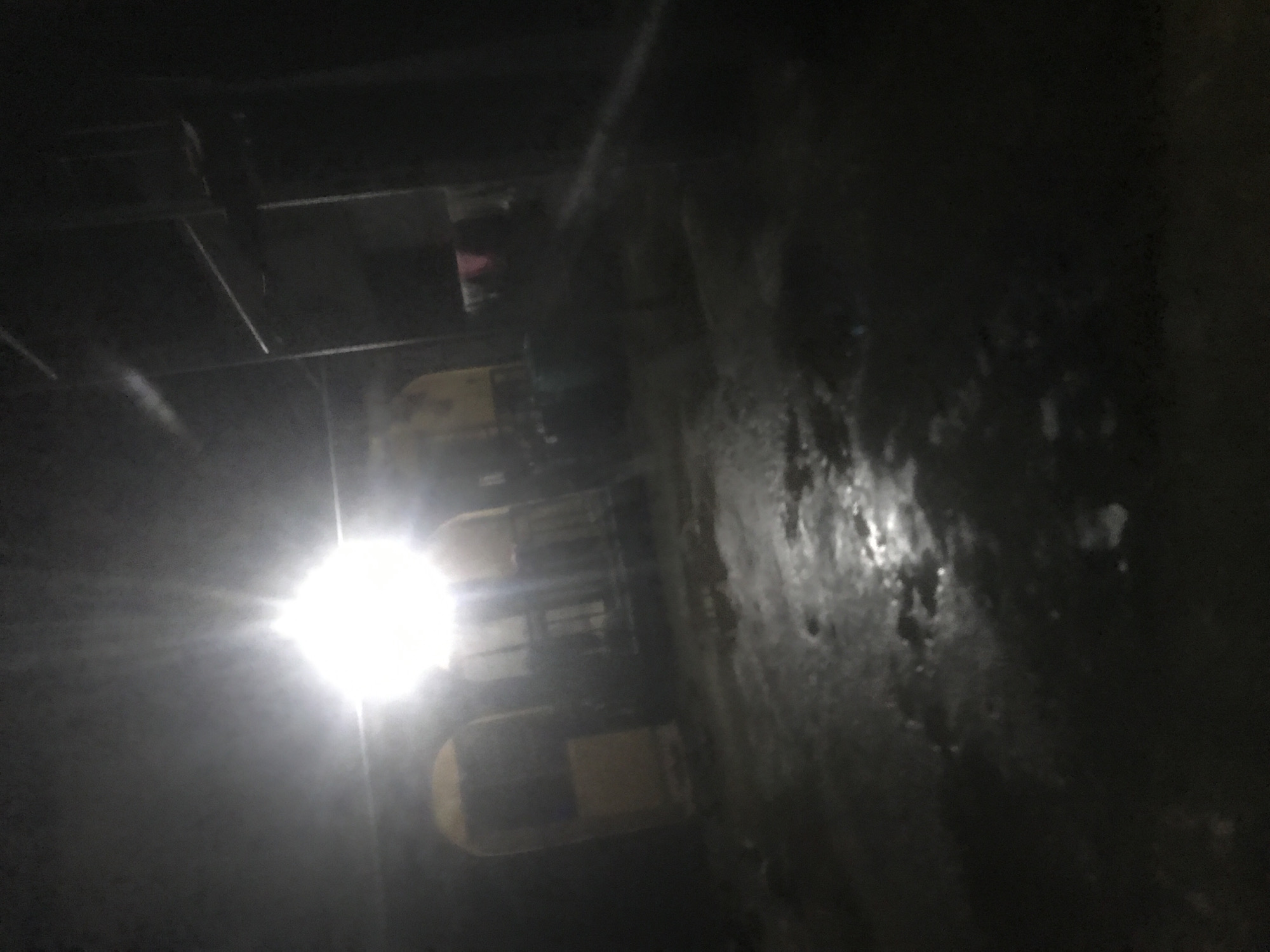 The image depicts a dark, possibly indoor, environment with some artificial lighting. A bright light source, likely a lamp, is prominent on the left side, casting strong glare and illuminating parts of the scene.
Several objects that appear to be yellow or light-colored containers or bins are visible, stacked or arranged in a row. Above these, there are some dark structures, possibly shelves or ceiling elements, and hints of other objects that are difficult to discern due to the low light conditions. In the upper central portion of the image, a small area of pink is visible, suggesting some fabric or material.
The scene appears to be somewhat industrial or utilitarian, given the nature of the objects and the lighting. There are no clear indications of weather or time of day, but the artificial light suggests it's not daytime or that the area is enclosed. The overall impression is one of darkness and limited visibility, with the bright light creating a stark contrast. There is no visible text in the image. The activity or event taking place is not clear from the visual information.