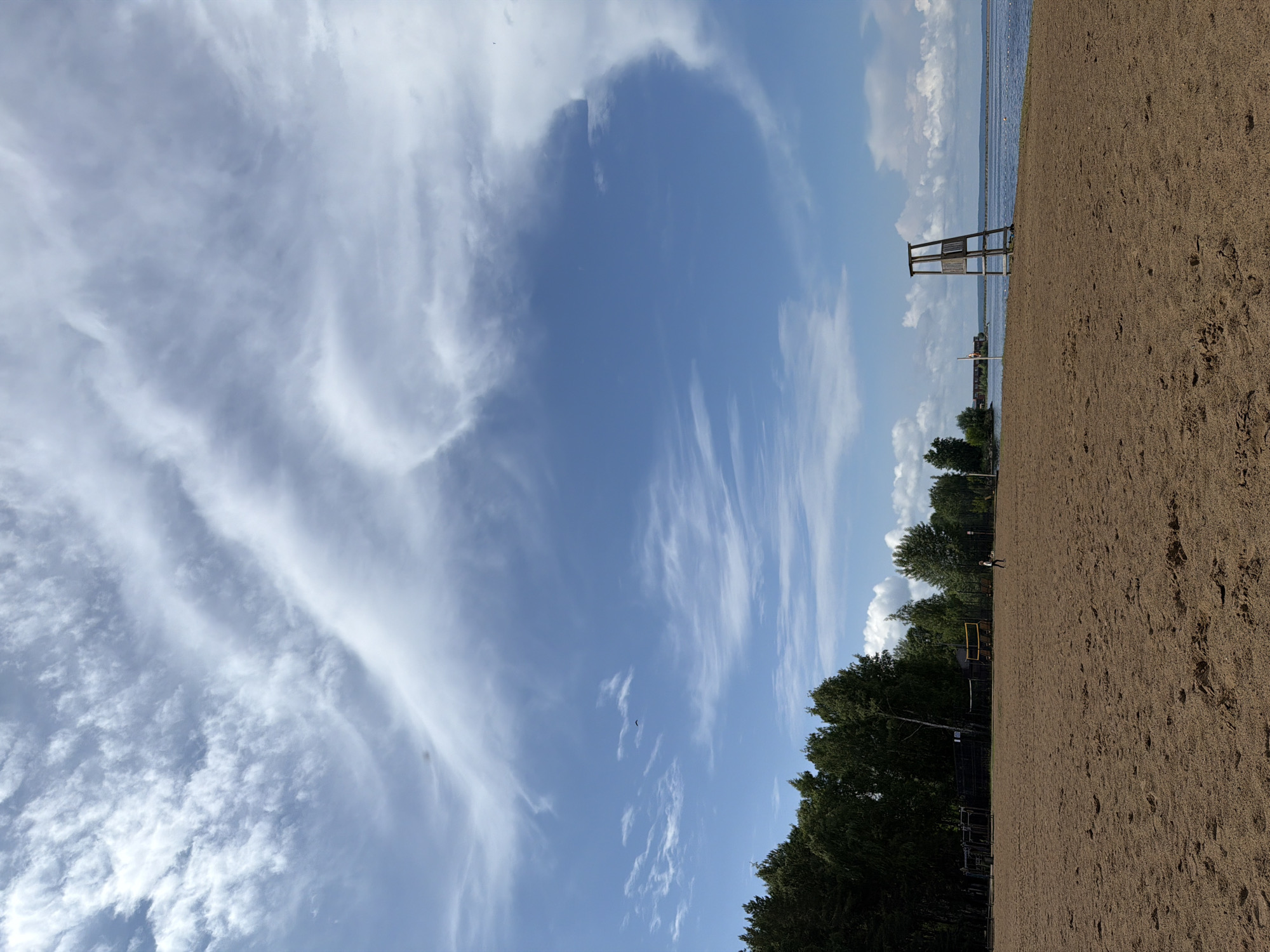 The image depicts a wide, sandy beach leading to a calm body of water under a vast sky filled with dramatic, wispy clouds. A wooden lifeguard tower stands prominently on the right side of the beach, overlooking the water. In the background, a line of dense green trees borders the far side of the beach, with a few volleyball nets visible amongst them, suggesting recreational activities. A single person can be seen walking in the distance on the sand. The overall scene is one of a serene, sun-drenched day at a coastal location. The specific location is on the Kama River embankment in Chaikovsky, Russia, a city known for its industrial significance and its proximity to the river.