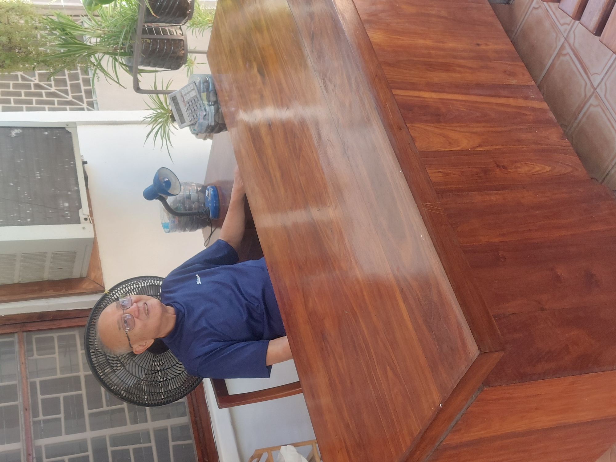An older man with glasses and short grey hair, wearing a blue short-sleeved collared shirt, is seated at a large, highly polished, dark reddish-brown wooden table or counter. He looks upwards and slightly to his left with a calm, slightly smiling expression. The wooden surface, possibly mahogany, shows prominent grain patterns and reflects light, indicating a smooth finish.
The setting appears to be an indoor or semi-enclosed space, likely a home or office in Dar es-Salaam, Tanzania. To the left of the man, a black oscillating pedestal fan is visible, and above it, a white wall-mounted air conditioning unit. A window with a white grid pattern is also seen in this area.
On the table behind the man, a small blue desk lamp, possibly LED, stands on a base made from a repurposed clear plastic bottle. Next to it, a grey calculator is also propped on what appears to be another repurposed plastic bottle. Further in the background, several green plants are arranged in suspended repurposed plastic bottles, adding a touch of greenery to the space. The wall behind the plants features a light-colored brick-like pattern. To the right of the table, a tiled floor or wall section with light brown geometric patterns is visible.
The bright lighting suggests it is daytime. The presence of an air conditioner and fan indicates a warm climate. No specific activity is clearly depicted, but the setup with a lamp and calculator suggests a work or study area. Text on the man's shirt is too blurry to decipher, though it appears to be a brand name or logo.
