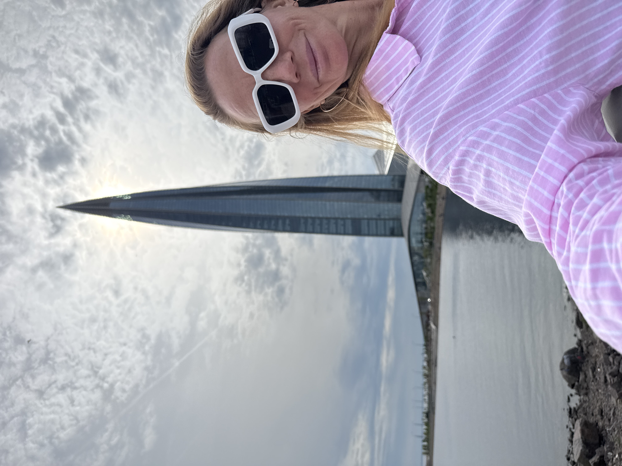 A woman wearing white-rimmed sunglasses and a pink-and-white striped button-down shirt takes a selfie outdoors. In the background is the Lakhta Center in Saint Petersburg, Russia, on a cloudy day. A body of water is visible to the right of the building. The ground appears to be rocky.
