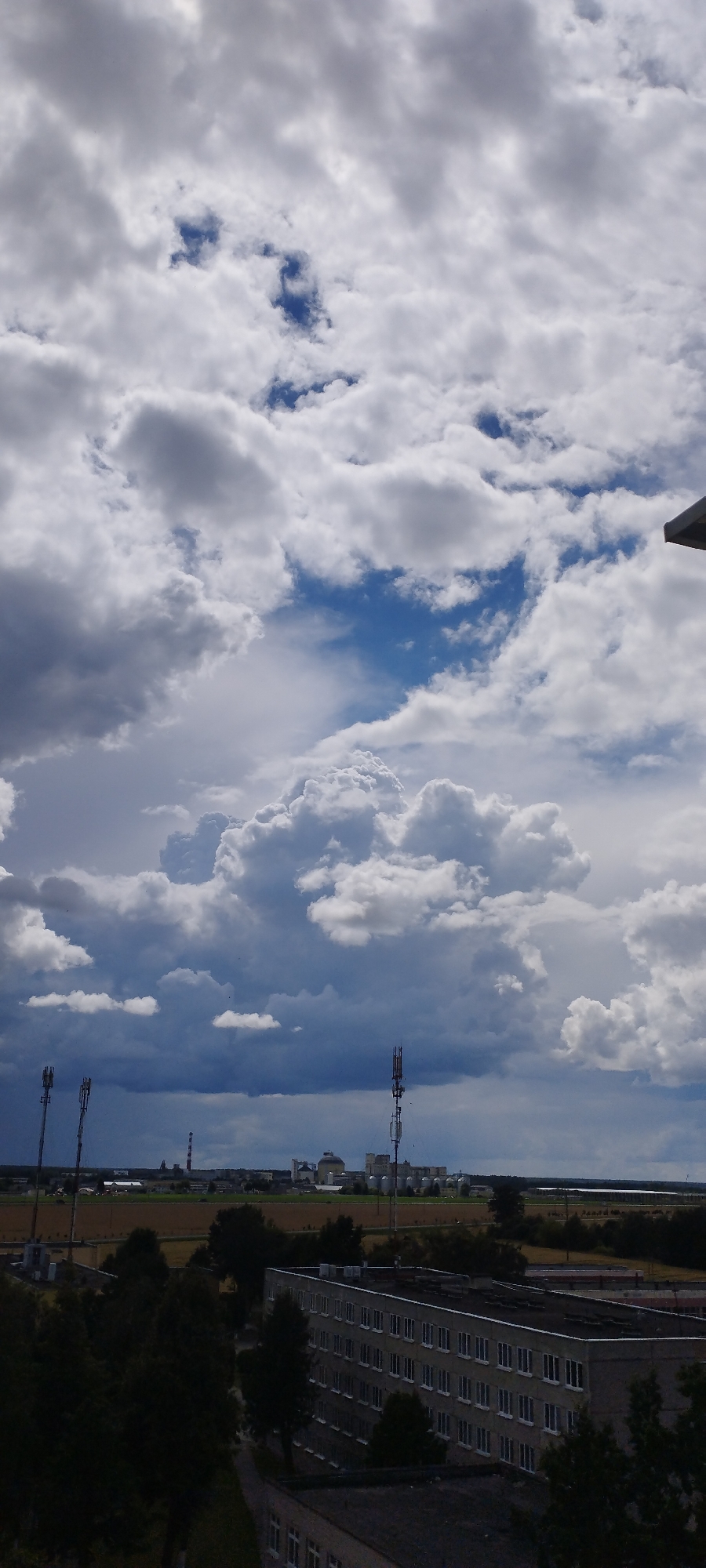 This is a photograph of a cloudy sky over the city of Skidzieĺ, Belarus. The image captures a dramatic sky filled with various cloud formations, ranging from light, wispy clouds to large, dark, thunderous clouds. Below the sky, a city landscape is visible, characterized by industrial buildings, agricultural fields, and communication towers.
In the foreground, the top of a building is visible, featuring a dark, flat roof structure. Behind it, a long, multi-story building with numerous windows is prominently featured. Trees and foliage are interspersed between the buildings and extend towards the horizon.
Further into the distance, a flat expanse of agricultural land is depicted. On this land, several industrial structures are present, including tall communication towers with antennas, and various other buildings, some with domed roofs. A tall, cylindrical smokestack is also visible, indicating industrial activity. The overall impression is one of a partly overcast day in an urban and industrial setting.