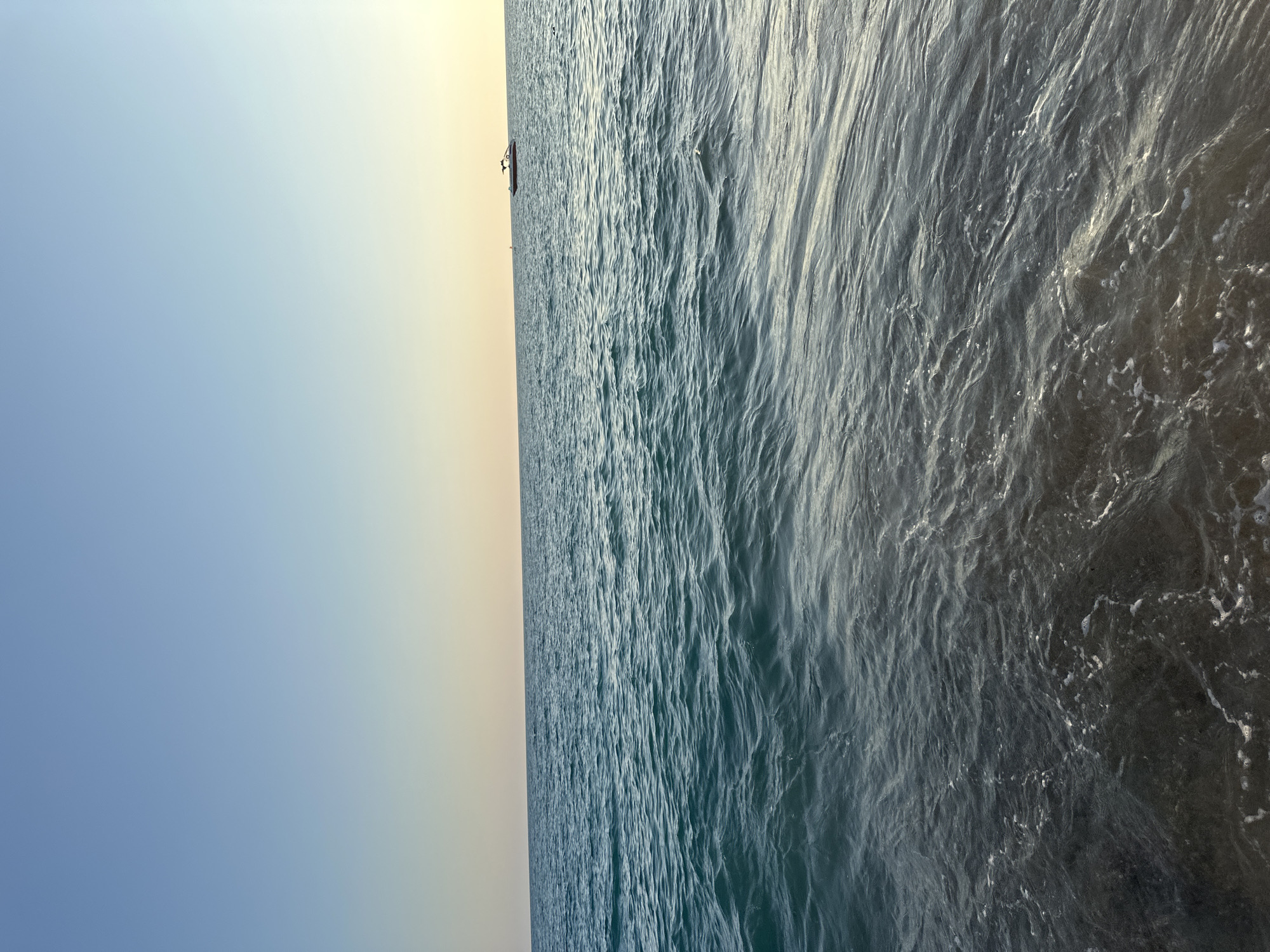 The image shows a body of water, likely the sea, under a clear sky with a gradient from blue at the top to a pale yellow near the horizon. The water has gentle waves. In the distance, a small red boat with a dark canopy is visible. The foreground shows the shallow water near the shore, with the seabed appearing dark and sandy, and some foam from the waves. The scene suggests a calm day at the coast, possibly during sunset or sunrise, given the sky's coloration. Based on the visual cues of a coastal setting and the presence of a boat, it is plausible that this image was taken at a beach along the coastline of Manavgat, Turkey, a region known for its Mediterranean beaches.