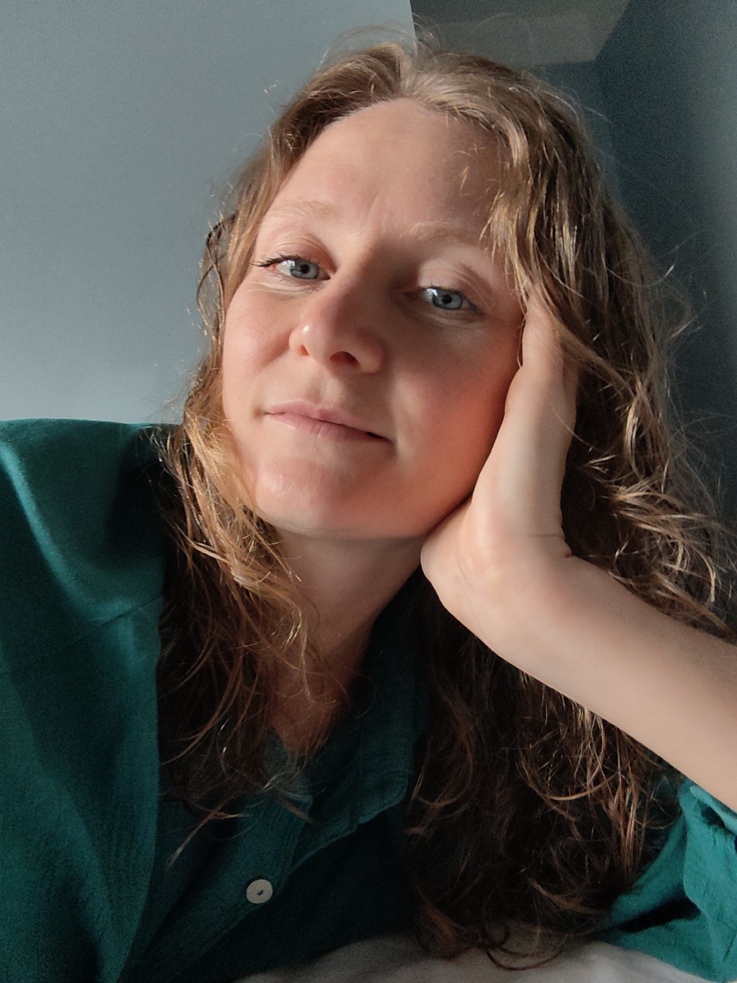 A woman with light skin, blue eyes, and wavy, shoulder-length blonde/light brown hair is looking directly at the camera. Her right hand is supporting her face, with her cheek resting against her hand. She is wearing a green button-up shirt. The background is a two-tone wall, with a lighter bluish-grey above and a darker grey below. The setting does not appear to be within Bai Chay Ward, Vietnam, or an urban environment.