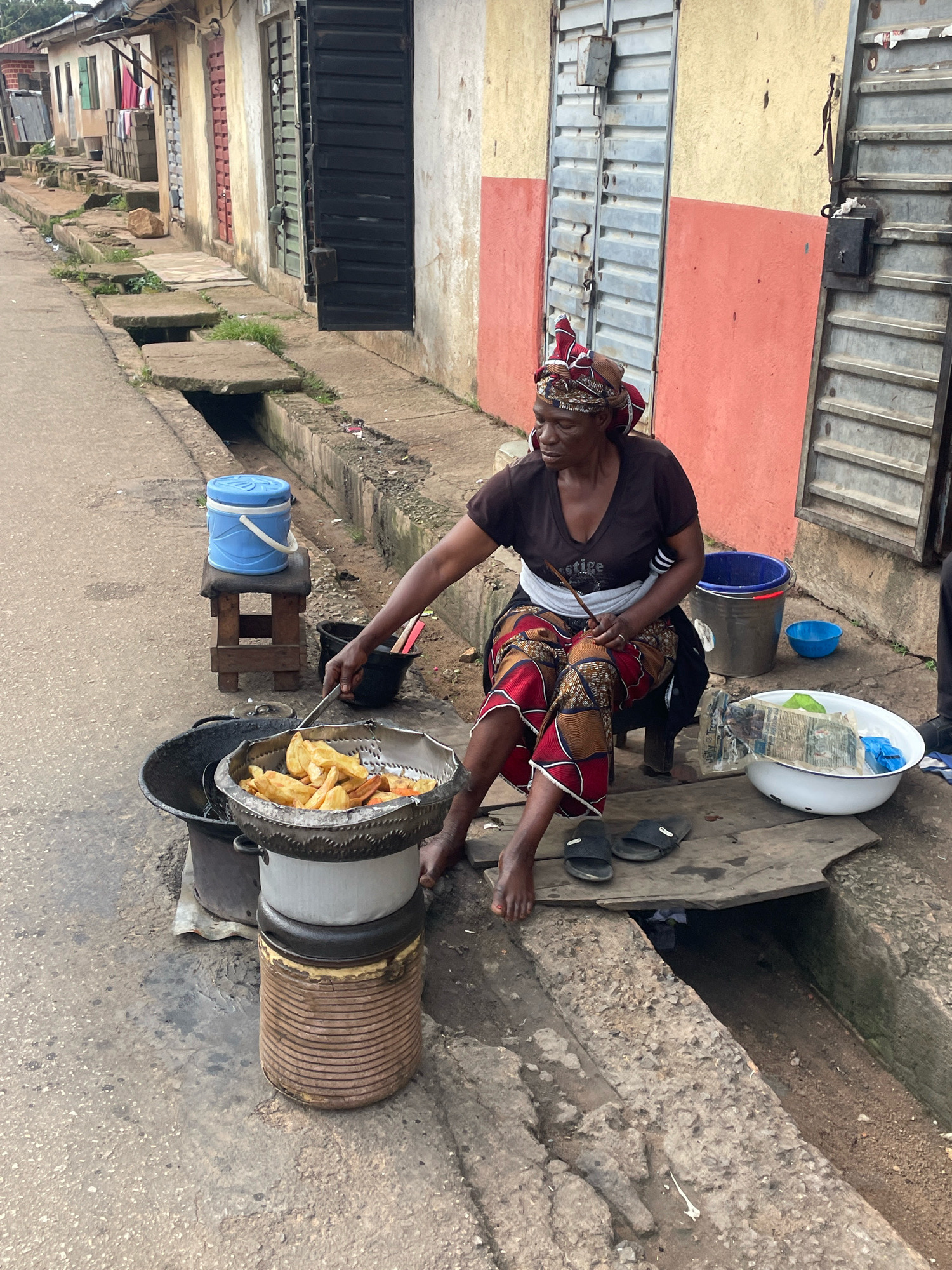 A woman is frying plantains in a street-side setting in Jos, Nigeria. She is wearing a headwrap and a patterned skirt, and she is seated on a stool next to a large pot of hot oil. A metal basket filled with fried plantains is suspended over the pot. Nearby, there is a blue water jug, several buckets, and a large white bowl containing more plantains and what appears to be plastic wrapping. The environment is a paved street with a concrete drainage ditch running alongside it. Buildings with metal shutters line the street, and various items are scattered on the ground. The overall impression is one of a bustling street food vendor.