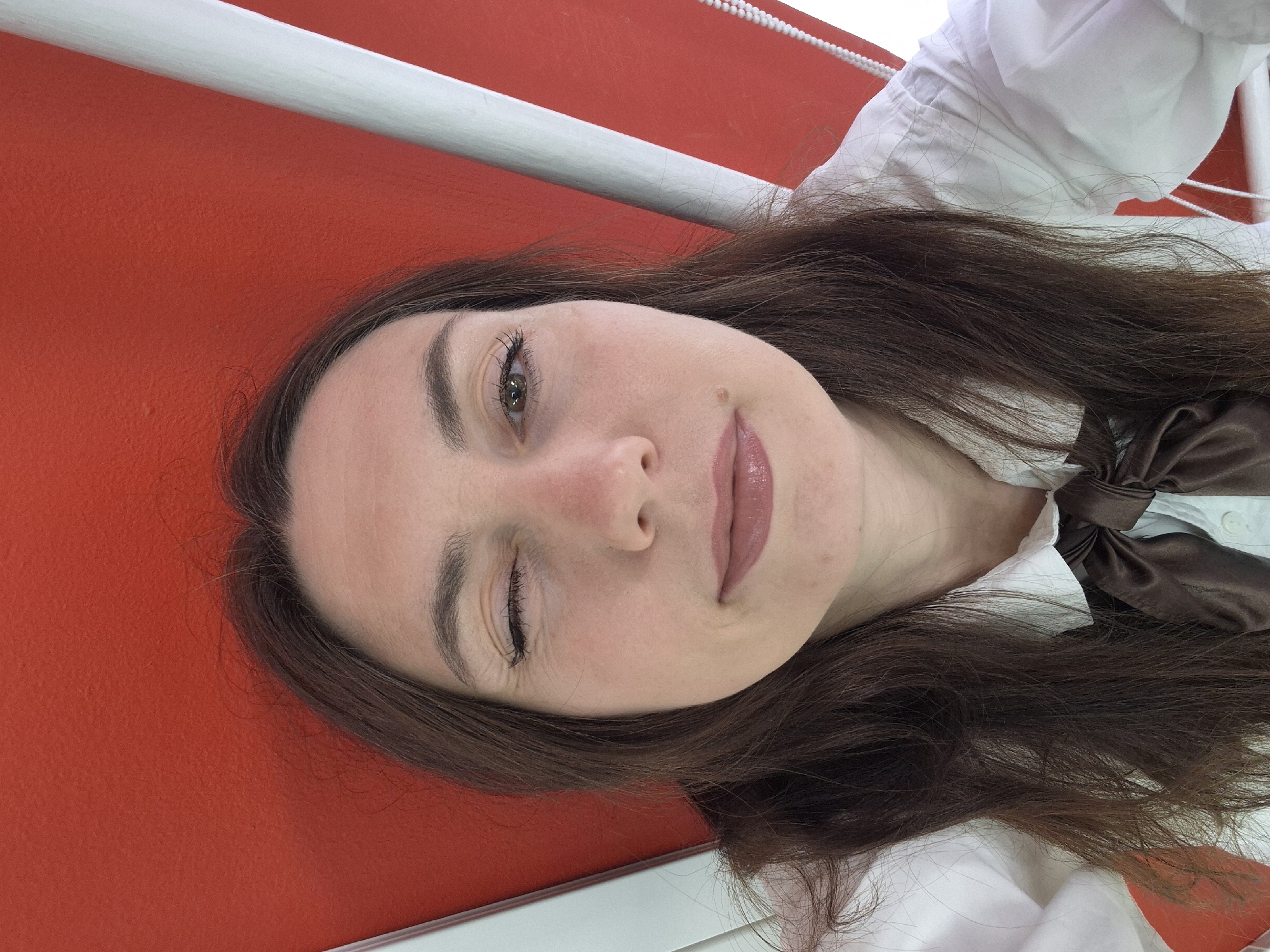 This close-up portrait, originally rotated 90 degrees clockwise, features a young adult woman with long, dark brown hair and fair skin. When viewed upright, her gaze is directed slightly to her left. Her left eye is open, revealing a light-colored iris, while her right eye is partly closed, suggesting a wink or a playful squint. Her lips are softly parted, adorned with light pink lipstick, and a small mole or beauty mark is visible on her right cheek. Her eyebrows are well-defined.
She is wearing a white collared shirt with a distinctive brown satin bow tied at the neck. Her dark hair flows around her, covering parts of her clothing.
The background consists of a vibrant, solid red surface, likely a wall or large piece of fabric. A white horizontal bar extends across the upper portion of the frame, and a white beaded string, resembling a blind pull cord, is partially visible in the upper right. These details suggest an indoor setting, possibly near a window equipped with blinds. The woman's expression appears relaxed and engaging, conveying a sense of playfulness. No text is visible in the image. The time of day and exact location are not discernible.