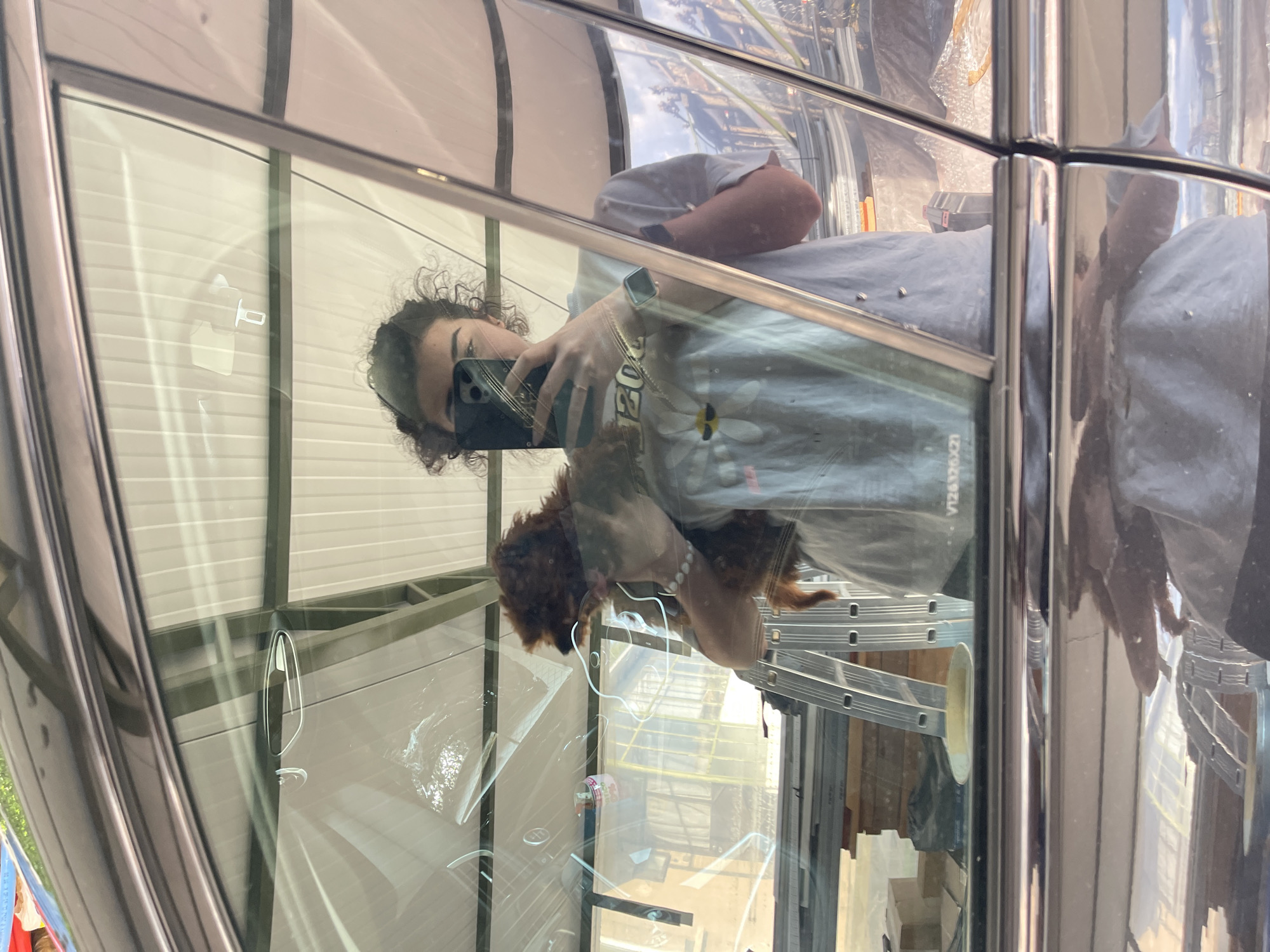 The image shows a reflection in a glass structure, possibly a building or a bus shelter. The reflection includes a person holding a mobile phone, presumably taking the picture, and a small, brown dog. The person is wearing a grey t-shirt with a flower design and the number "2021". Behind the person and dog in the reflection is a ladder and construction materials. The image was taken in Sofia, Bulgaria.