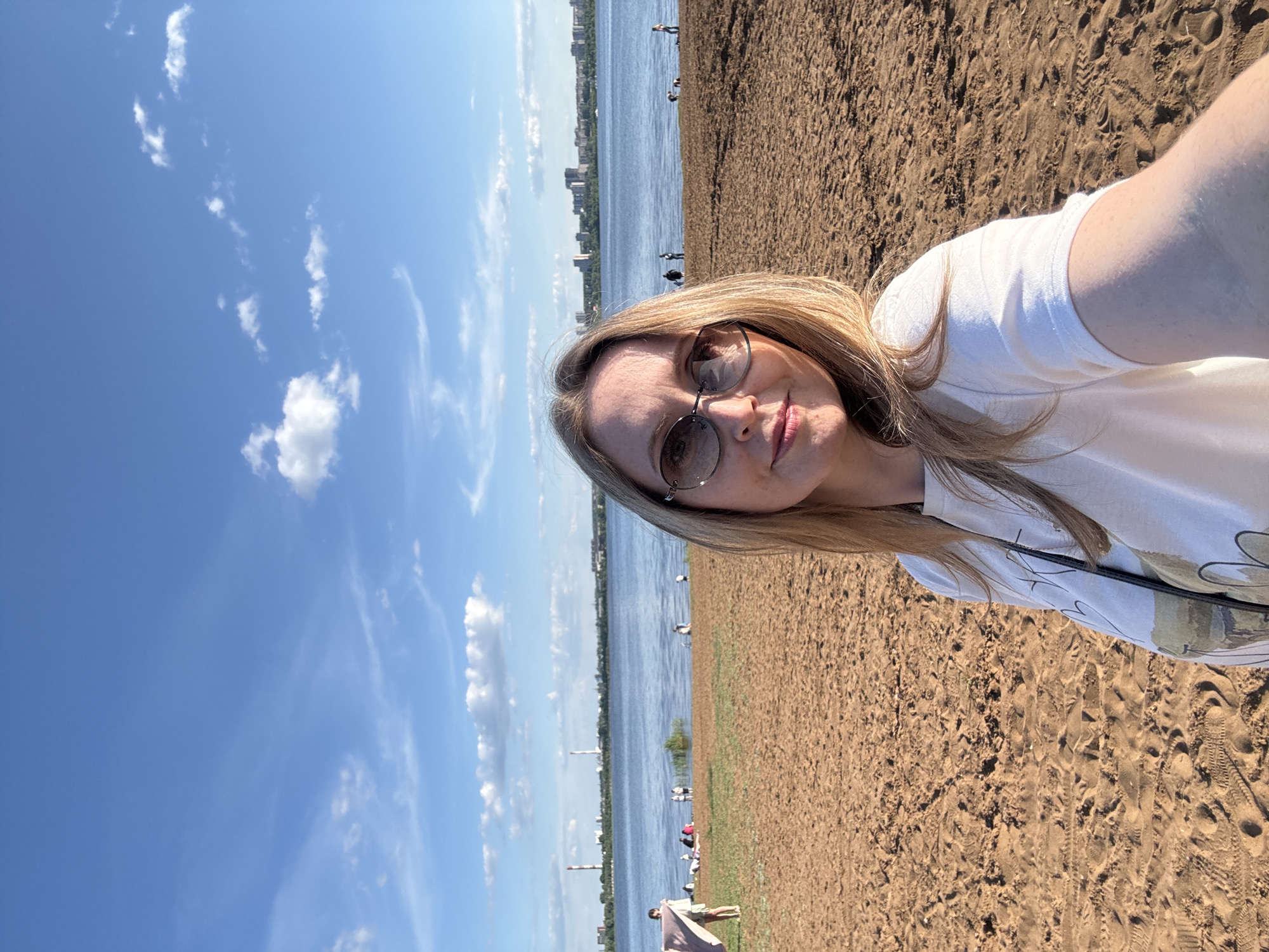 An eye-level selfie captures a woman with blonde hair and dark, oval-framed sunglasses, wearing a white short-sleeved t-shirt with a subtle graphic design on the left chest. A dark strap, possibly from a bag, crosses her left shoulder. She exhibits a slight smile while holding the camera. The foreground consists of a wide, light reddish-brown sandy beach covered with numerous footprints. Beyond the beach, the calm, blue waters of the Izhevsk Pond extend, reflecting the sky. Several individuals are visible on the beach and in the shallow water; one person on the far left stands holding a light-colored blanket. The distant background reveals the cityscape of Izhevsk, Russia, with a skyline of numerous buildings interspersed with green trees. On the far left of the distant shore, two prominent, tall, red and white striped industrial smokestacks, characteristic of the Izhevsk Thermal Power Plant (Izhevskaya CHPP-1), rise against the horizon. The sky is clear blue with scattered white cumulus and wispy cirrus clouds. The scene is illuminated by direct sunlight.