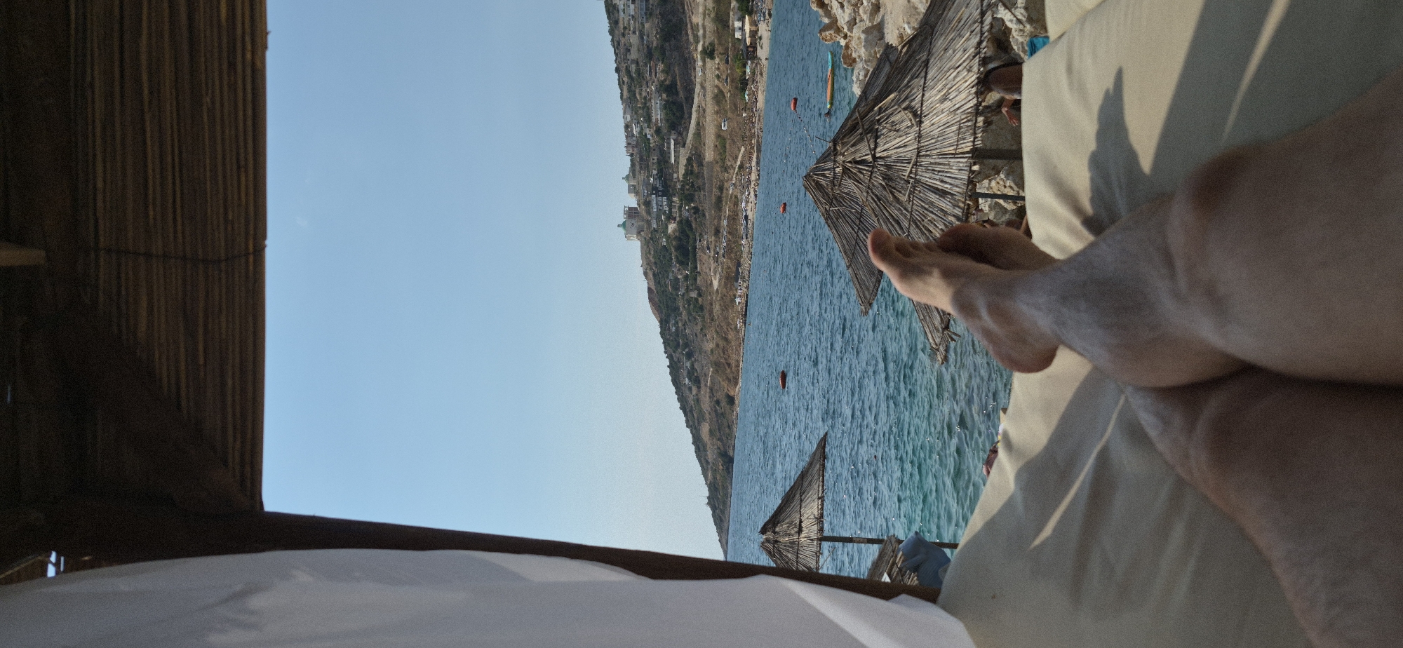 The image depicts a first-person perspective from a lounge chair under a thatched-roof canopy, offering a view of a beach and the sea. In the foreground, a person's legs and feet are visible, resting on a beige cushion. The sea is a vibrant blue, with gentle waves. To the left, a white curtain is draped from the canopy. Two thatched umbrellas are present on the beach, one closer and one further away. In the middle ground, there are rocky areas and more beach umbrellas. The background features a hillside dotted with buildings and vegetation, characteristic of the city of Saranda, Albania. The sky is clear and blue. Several orange buoys are visible in the water. A few people are subtly visible in the background, enjoying the beach.