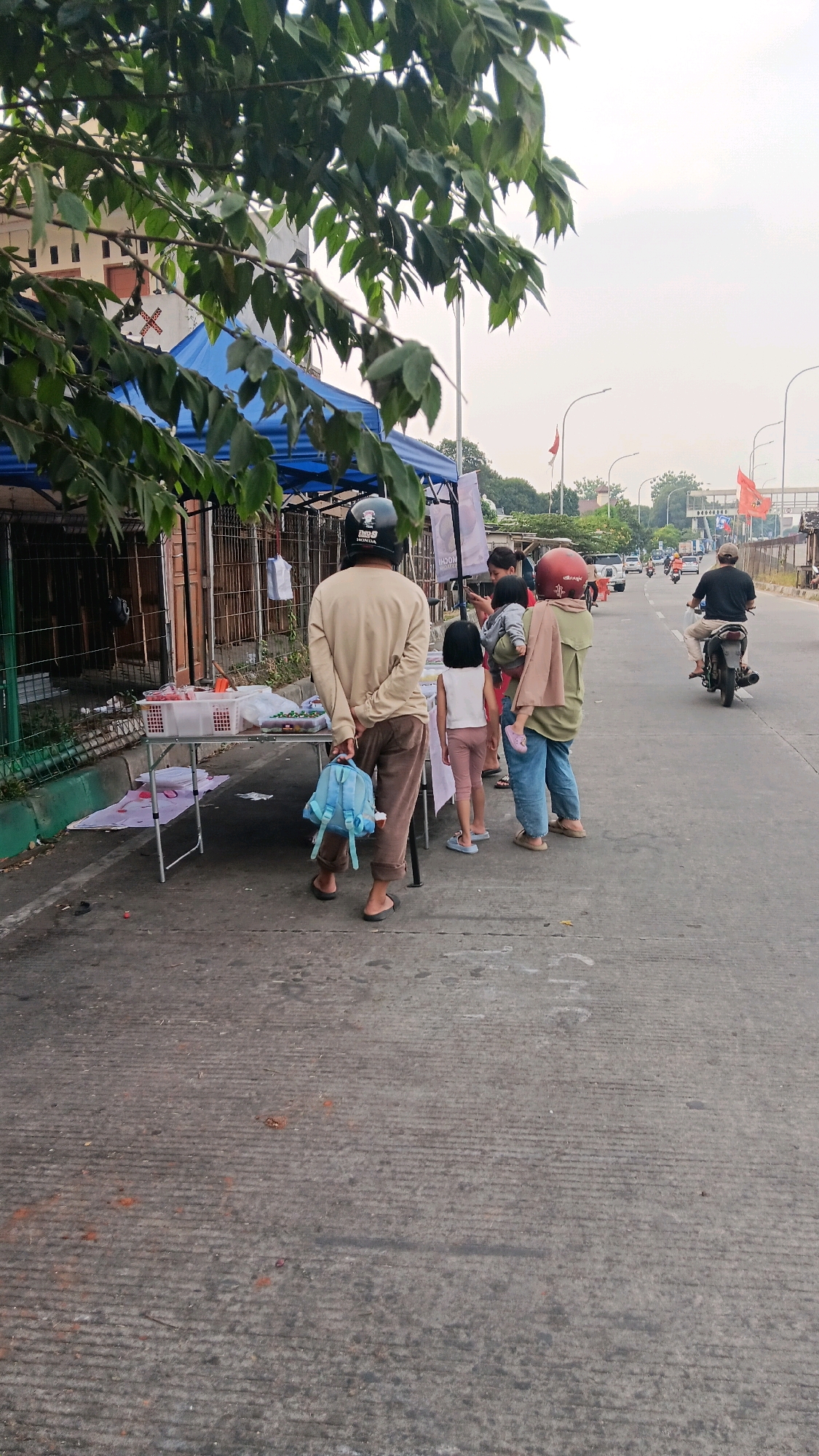 The image depicts a street scene in Depok, Indonesia, with a family of four, including two adults and two children, standing near a makeshift stall under a blue tent. The stall displays various items in white baskets. The family appears to be waiting or observing. A man wearing a helmet and a light-colored long-sleeved shirt is holding a small blue backpack. A woman, also wearing a helmet and a green top, is holding a baby. A young girl in a white top and pink pants stands near them. In the background, a man on a motorcycle is seen riding down the street, and there are other vehicles and buildings visible. The environment appears to be a roadside in a moderately busy urban area.