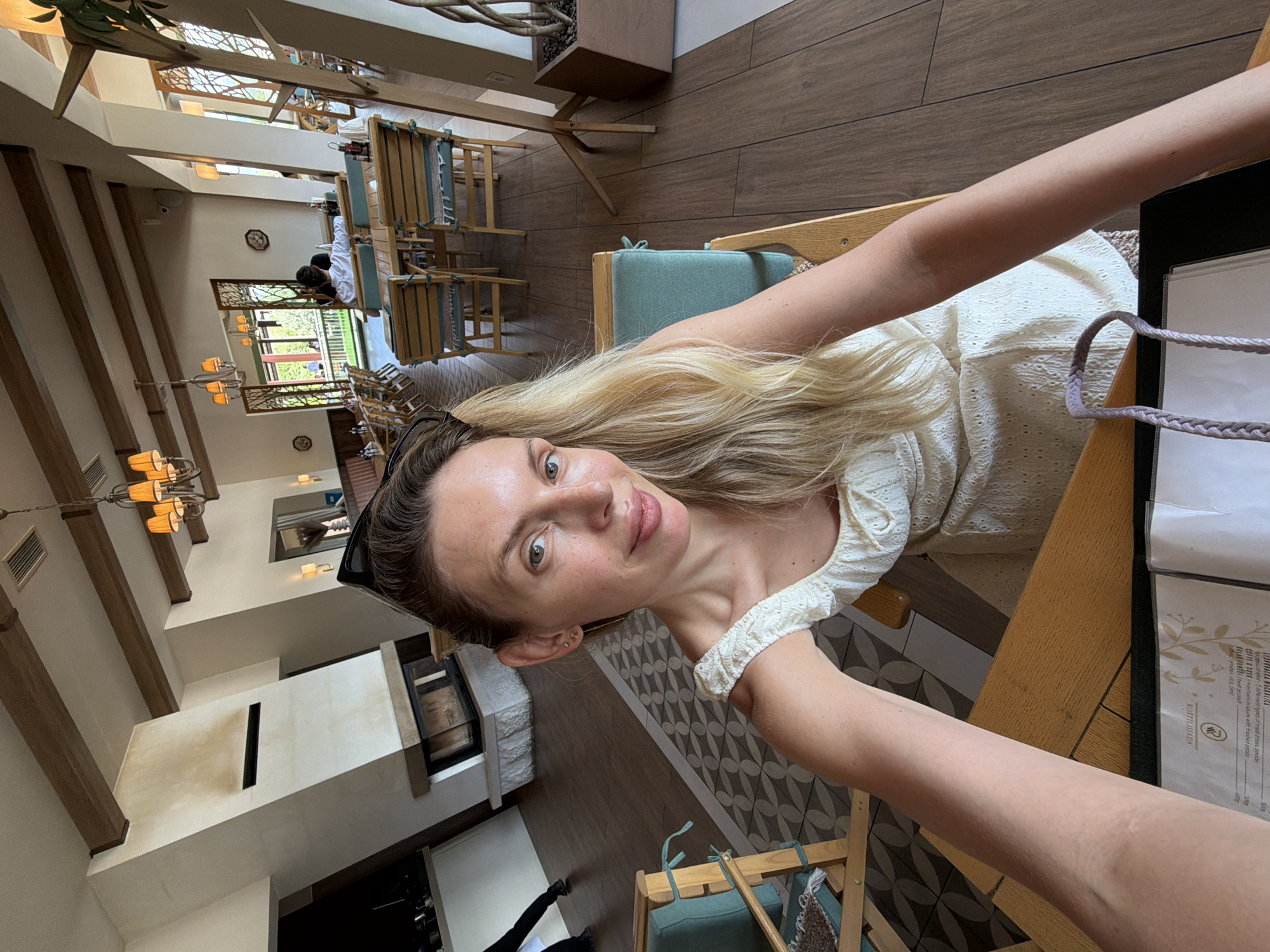 A woman with blonde hair is taking a selfie in what appears to be a restaurant or cafe. She is wearing a white, textured dress. Her arm is extended towards the camera, and she is smiling. She has sunglasses perched on her head.

The setting is a spacious interior with wooden beams on the ceiling and a wooden floor. There are tables and chairs with green cushions. In the background, there are decorative wooden screens and pendant lights. Further in the background, there are glimpses of other patrons and a fireplace. The overall ambiance suggests a relaxed daytime setting.

The visible text on a menu card on the table reads "ROSTILJIJADA" and mentions types of meat and side dishes.