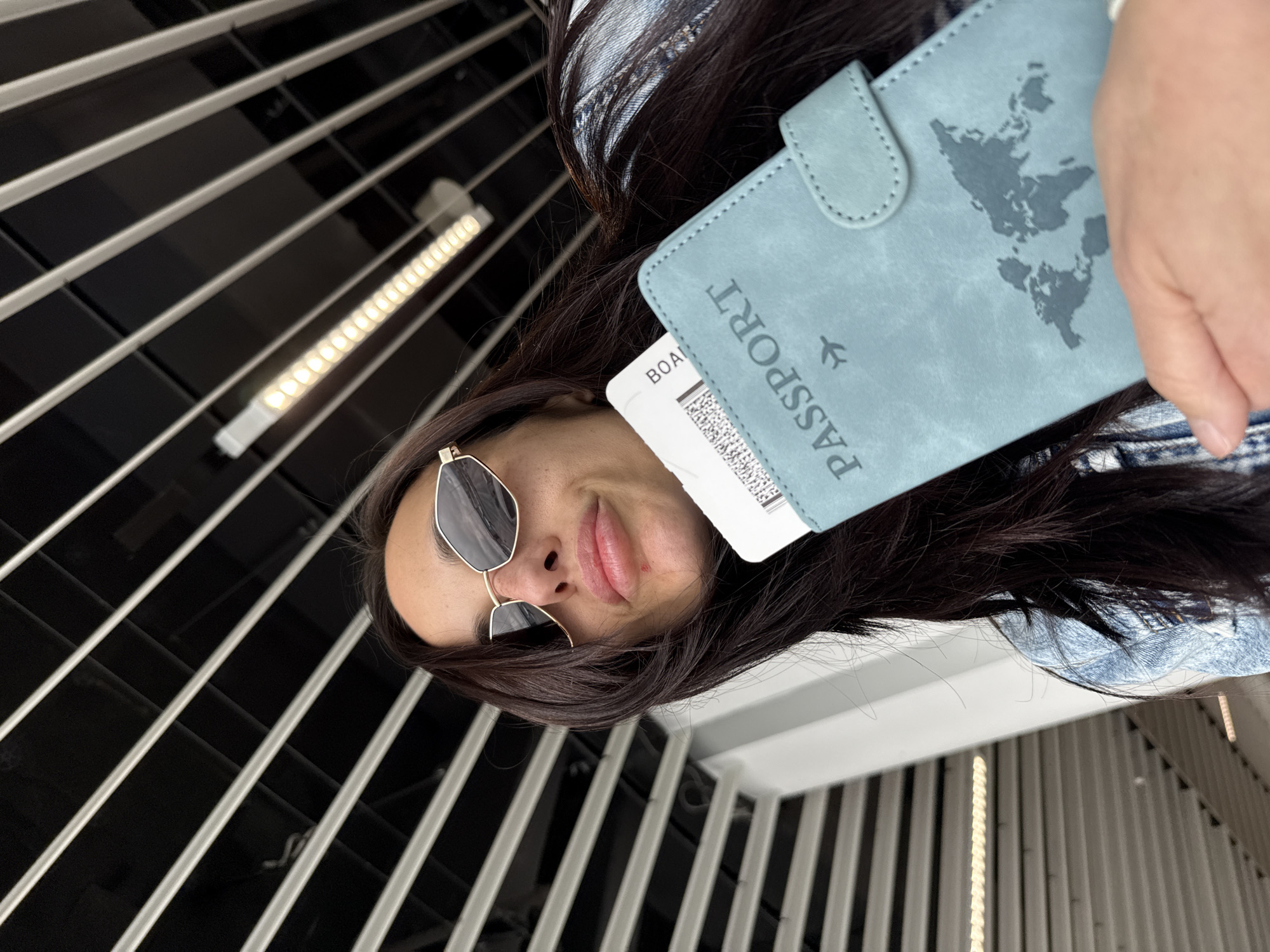 A woman wearing sunglasses holds a light blue passport and a boarding pass. The background shows an architectural structure with horizontal and vertical lines. The location cannot be confirmed as Sofia, Bulgaria without additional context or landmarks.