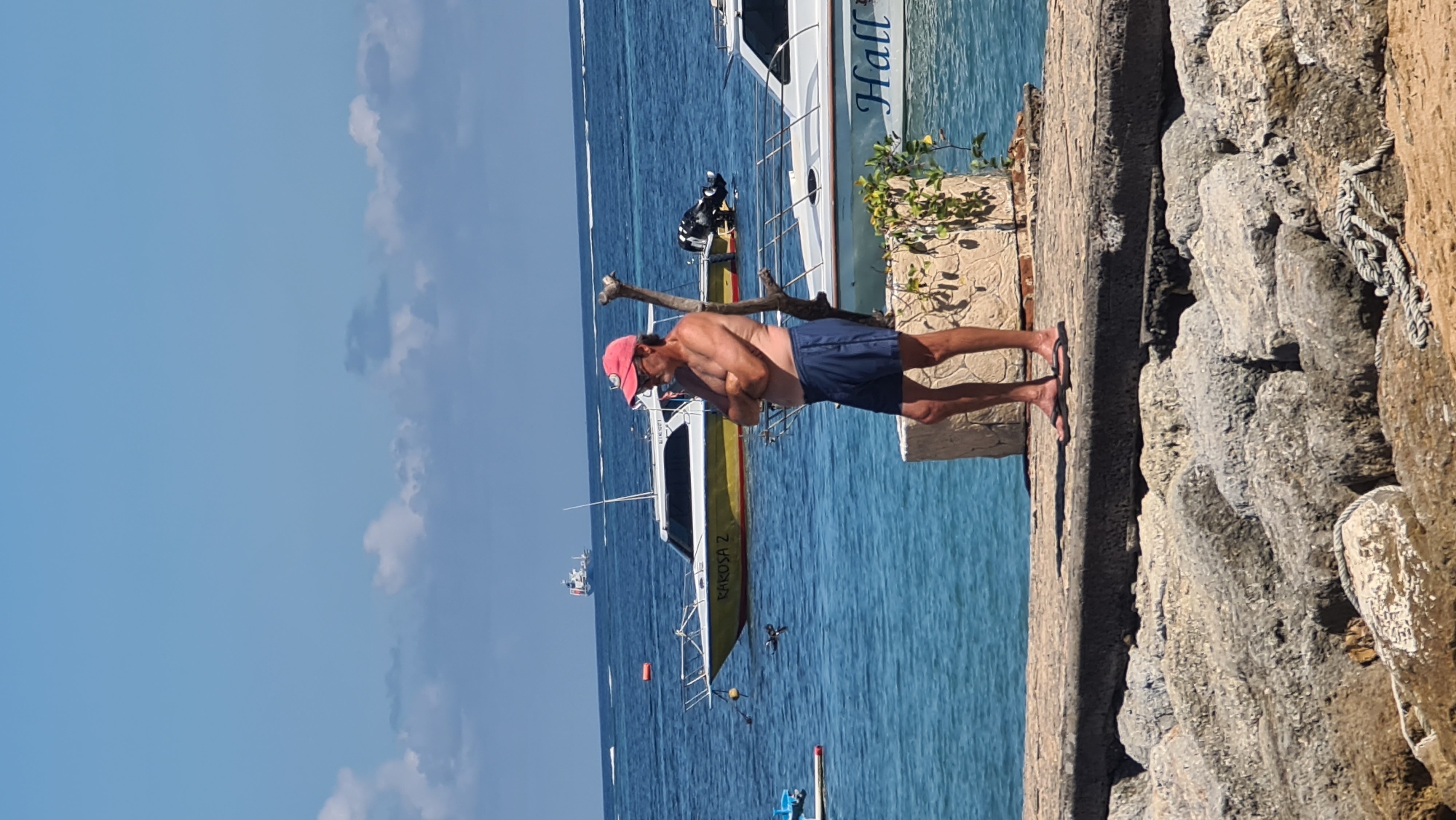 A man wearing a pink baseball cap, sunglasses, and blue shorts stands on a concrete pier with his arms crossed. He is shirtless and has a tan. Behind him, the turquoise ocean is visible with several boats. One yellow and white boat is labeled "RAROSA 2". Another white boat with "Hall" written on its side is closer to the pier. In the distance, a larger ship can be seen on the horizon. The sky is clear blue with a few scattered clouds. The pier is constructed with rough, gray rocks and concrete slabs. The scene appears to be a coastal area in or near Denpasar, Indonesia, likely a public beach or harbor.