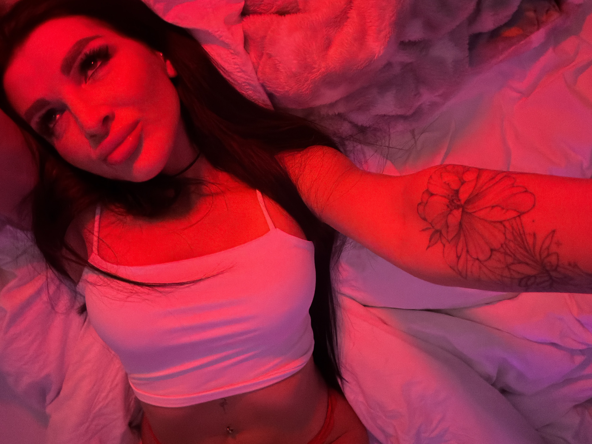 A person with long, dark hair is lying on a bed covered with white blankets and a fluffy pink and purple blanket. They are wearing a white tank top. There is a floral tattoo on their arm. The image is lit with red light. There is no evidence to place this image in the city of Boreti, Montenegro.