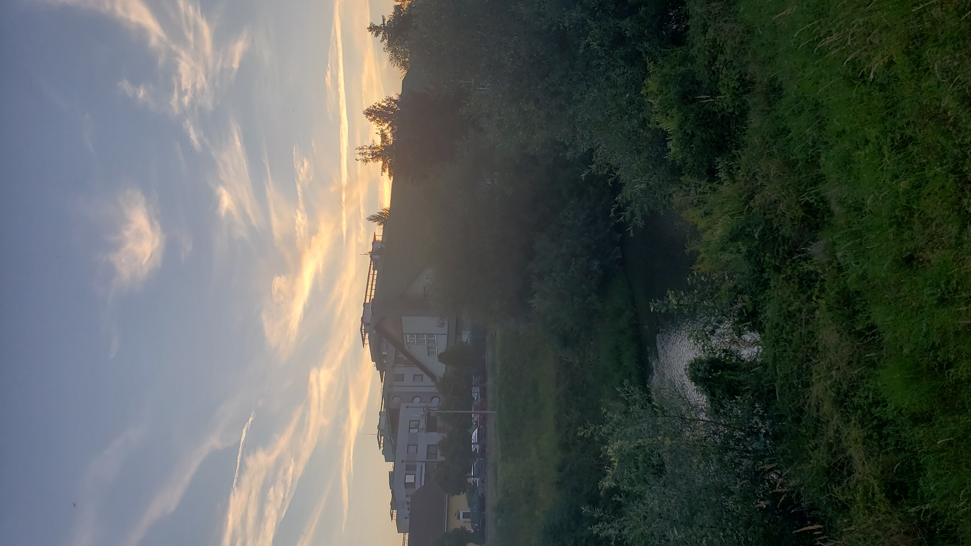 The image depicts a serene sunset scene in Prešov, Slovakia, likely near the Torysa River. In the foreground, lush green grass and dense bushes dominate, creating a natural buffer. Beyond the foliage, a calm body of water reflects the soft hues of the twilight sky, with some ripples suggesting gentle movement.
Across the water, a road is visible, lined with parked cars. Behind the road, a series of residential buildings are situated, ranging from lower-rise structures to taller apartment blocks. The buildings appear to be modern, with flat roofs and multiple stories. Trees are interspersed between the buildings and along the roadside, contributing to the urban landscape.
The sky above is a canvas of soft blues and oranges, with wispy clouds catching the last rays of the setting sun. The overall atmosphere is peaceful and tranquil, capturing the end of a day in this Slovakian city. There are no people or animals visible in the image, making it a landscape-focused portrayal of the environment.