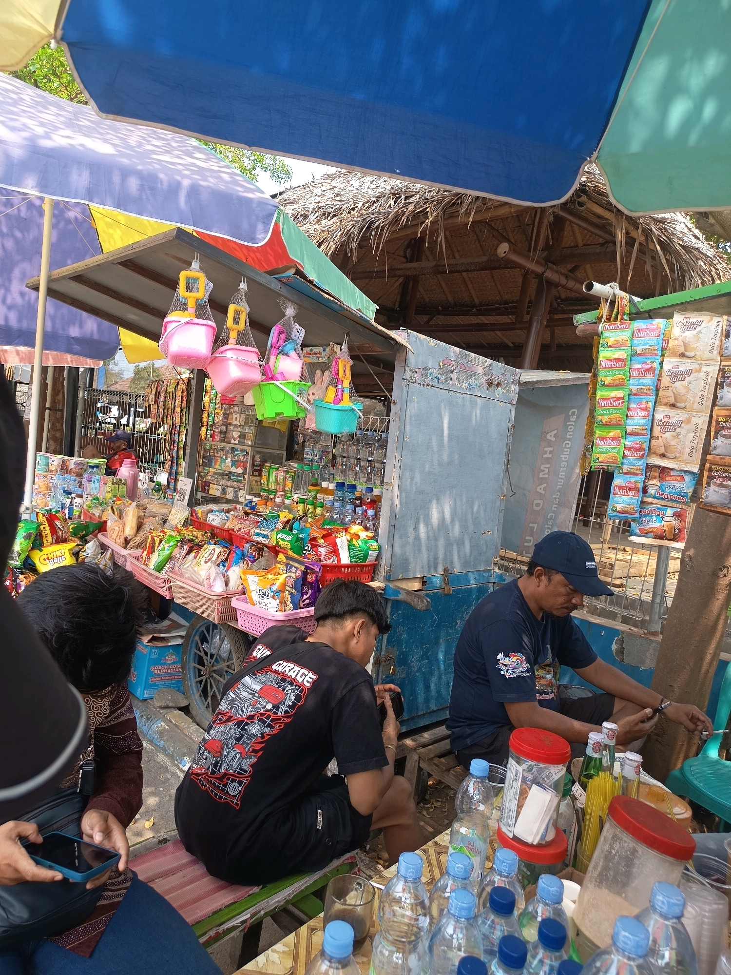 The image depicts a vibrant street-side stall in Jepara, Indonesia, bathed in bright sunlight. The stall is covered by a multi-colored umbrella and a thatched roof structure, suggesting an outdoor setting, possibly near a beach or tourist area given the presence of colorful plastic buckets and spades hanging from the stall.
The stall itself is laden with a variety of goods, primarily packaged snacks and drinks. Baskets overflowing with chips, biscuits, and other treats are arranged on a cart. Numerous bottles of water and other beverages are neatly stacked. Several packets of coffee and powdered drinks hang from a display rack on the right side of the stall.
Three individuals are visible in the scene. In the foreground, two young men are seated on a bench in front of the stall. One is wearing a black t-shirt with a graphic design on the back and appears to be holding a phone or camera, possibly taking a picture. The other is partially obscured, but also appears to be looking towards the stall. To the right of the stall, an older man wearing a blue t-shirt and a dark blue cap is seated, looking down and appearing to be engaged with his phone or perhaps a purchase.
The overall atmosphere is that of a busy, casual marketplace, where locals and possibly tourists can purchase refreshments and snacks. The presence of the umbrellas and the open-air setup strongly indicates a warm, tropical environment. The colorful displays and abundance of products suggest a well-stocked and popular local vendor.